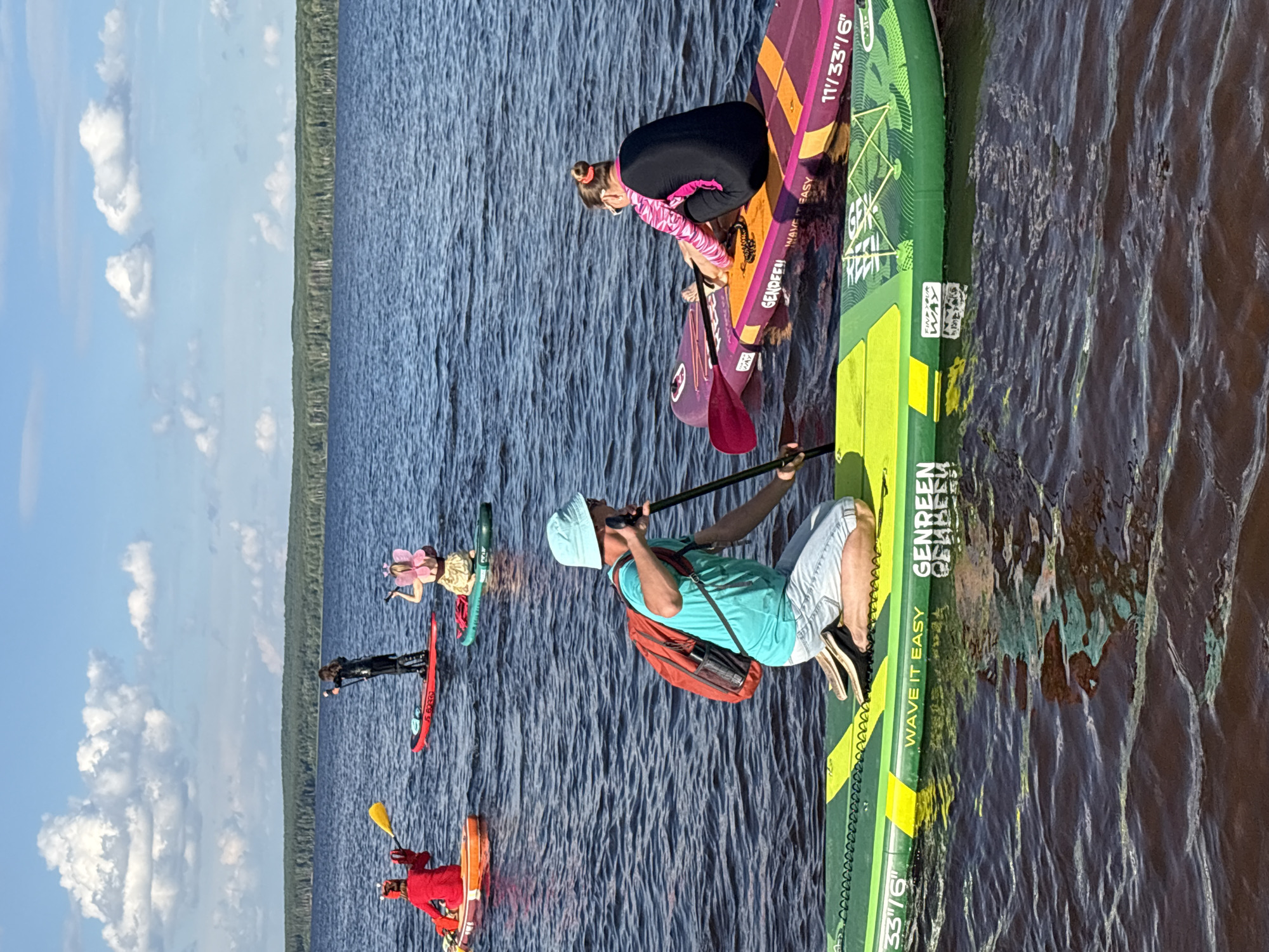 On a wide, dark blue body of water, under a partly cloudy blue sky, six people are paddleboarding. In the foreground, a man in a light blue bucket hat, turquoise t-shirt, and light-colored shorts kneels on a green and yellow paddleboard, paddling with a black and pink paddle. Behind him to the right, a woman in a black and pink wetsuit sits on a purple and orange paddleboard, facing away from the camera. Further back, three people are visible: a person in a black outfit standing on a red paddleboard, a person in a red costume on an orange and white paddleboard, and a person in a tan costume with pink wings on a green paddleboard. The paddleboarders are spread out across the water, with a dense, green forest line visible in the distance. This scene likely takes place on the Votkinsk Reservoir, a man-made lake located on the Kama River, near the city of Chaikovsky, Russia.