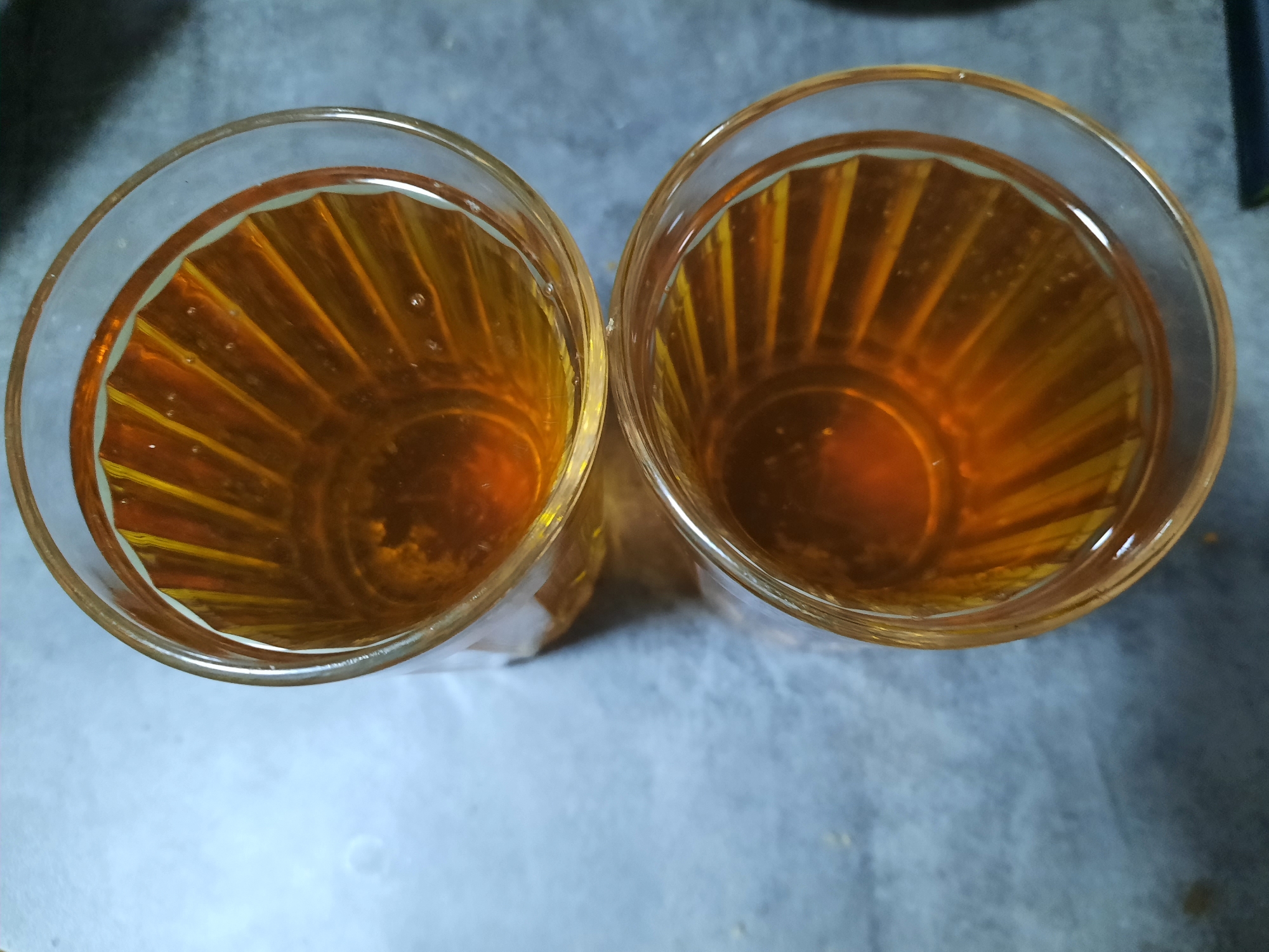 The scene depicts two clear glass tumblers, viewed from directly above, placed on a light gray, subtly textured surface. Both tumblers are filled with an amber-colored liquid, presenting a transparent appearance. The inner surface of each glass features a vertical ribbed or fluted design, creating distinct light and shadow patterns within the liquid. Numerous small, translucent bubbles are visible within the liquid, particularly near the surface in both glasses. No individuals, actions, or interactions are observable. The exact location within the city of Krasnogorskoye, Russia, cannot be determined from the visual evidence presented.