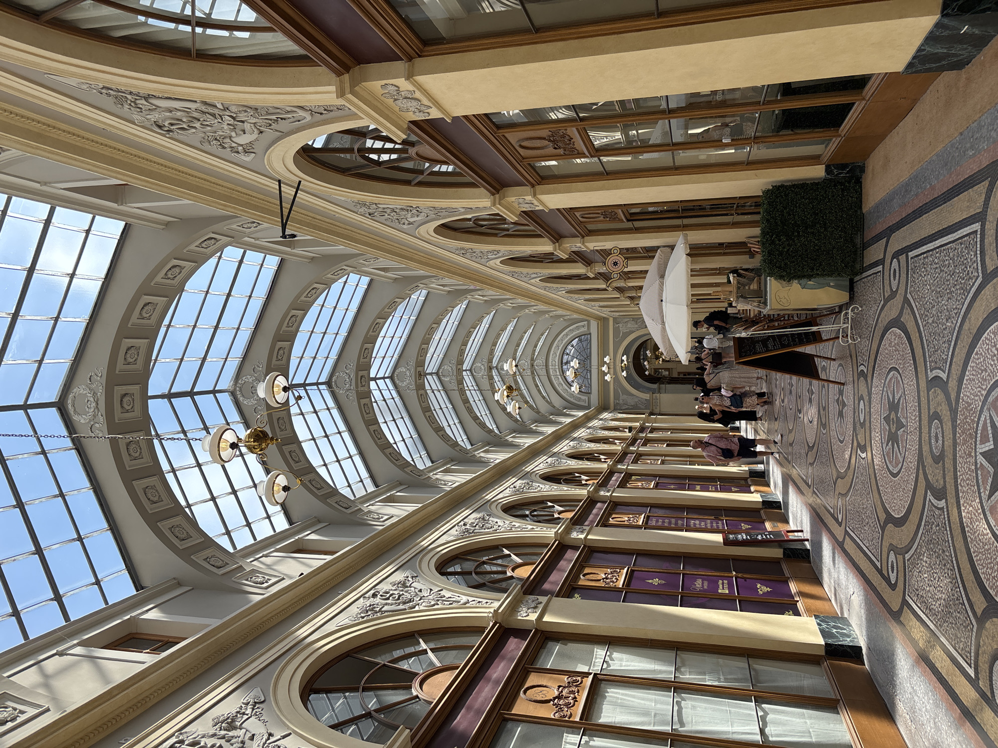 This is an interior shot of the Galerie Vivienne in Paris, France, a covered passage lined with shops and cafes. The ornate architecture features a high, arched glass ceiling that allows natural light to flood the space, illuminating the detailed mosaic tile flooring. Shops with large display windows and decorative facades line both sides of the passage. Several people are seen walking through the passage, some browsing shop windows, while others are seated at tables near a cafe area. The scene depicts a bustling, yet elegant, Parisian landmark on a bright, sunny day.