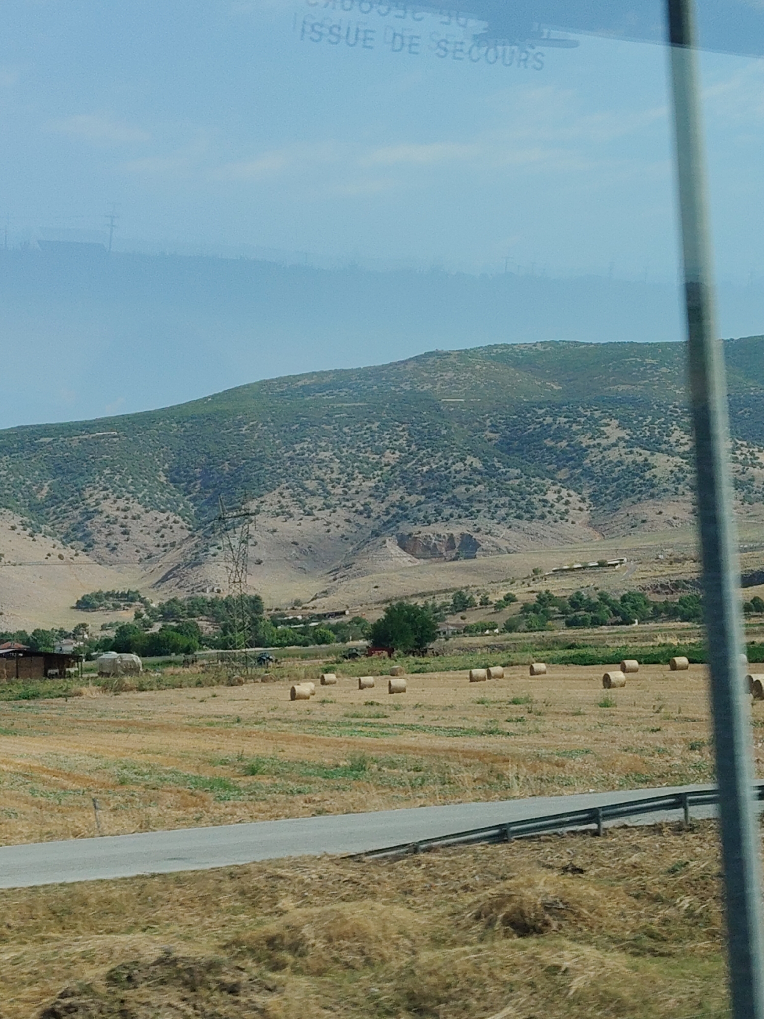 A wide shot captures a rural landscape beneath a hazy sky, with a rocky, scrub-covered mountain dominating the background. In the mid-ground, a field of dry, golden grass is dotted with numerous cylindrical hay bales. A narrow, grey asphalt road with a metal guardrail cuts across the lower portion of the frame. A tall electrical pylon stands prominently in the middle distance, to the left of the mountain's center. To the left of the pylon, a small cluster of buildings with red roofs is visible, adjacent to a stack of straw bales. Further to the left, a patch of green trees provides a contrast to the dry vegetation. The overall scene depicts a sun-drenched, agricultural setting in Taxiarches, Greece.