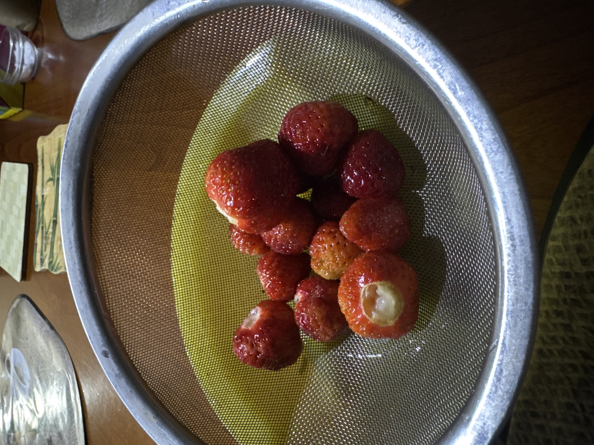 An overhead photograph depicts a metal mesh colander holding approximately 12-15 red strawberries, positioned in an indoor setting within Odesa, Ukraine. The circular colander features a fine mesh screen and a prominent, worn silver-colored metal rim. Beneath the colander, a bright yellow bowl or plate is visible, intended to collect any liquid. The strawberries exhibit varying shades of red, indicative of ripeness; one large strawberry in the foreground has a distinct, clean circular cavity, suggesting it has been hulled, and another further back displays a similar indentation. Their surfaces appear glistening and wet, consistent with recent washing. The colander rests on a polished wooden table. In the upper-left background, a reflective, possibly metallic or glass, oval-shaped coaster or plate is partially visible. The upper-right background reveals a small, clear glass bowl containing a white granular substance, likely salt or sugar, alongside a segment of a light-colored placemat adorned with a green bamboo-leaf pattern. Further back, a portion of a colorful package or container is faintly discernible. No individuals are present in the frame.