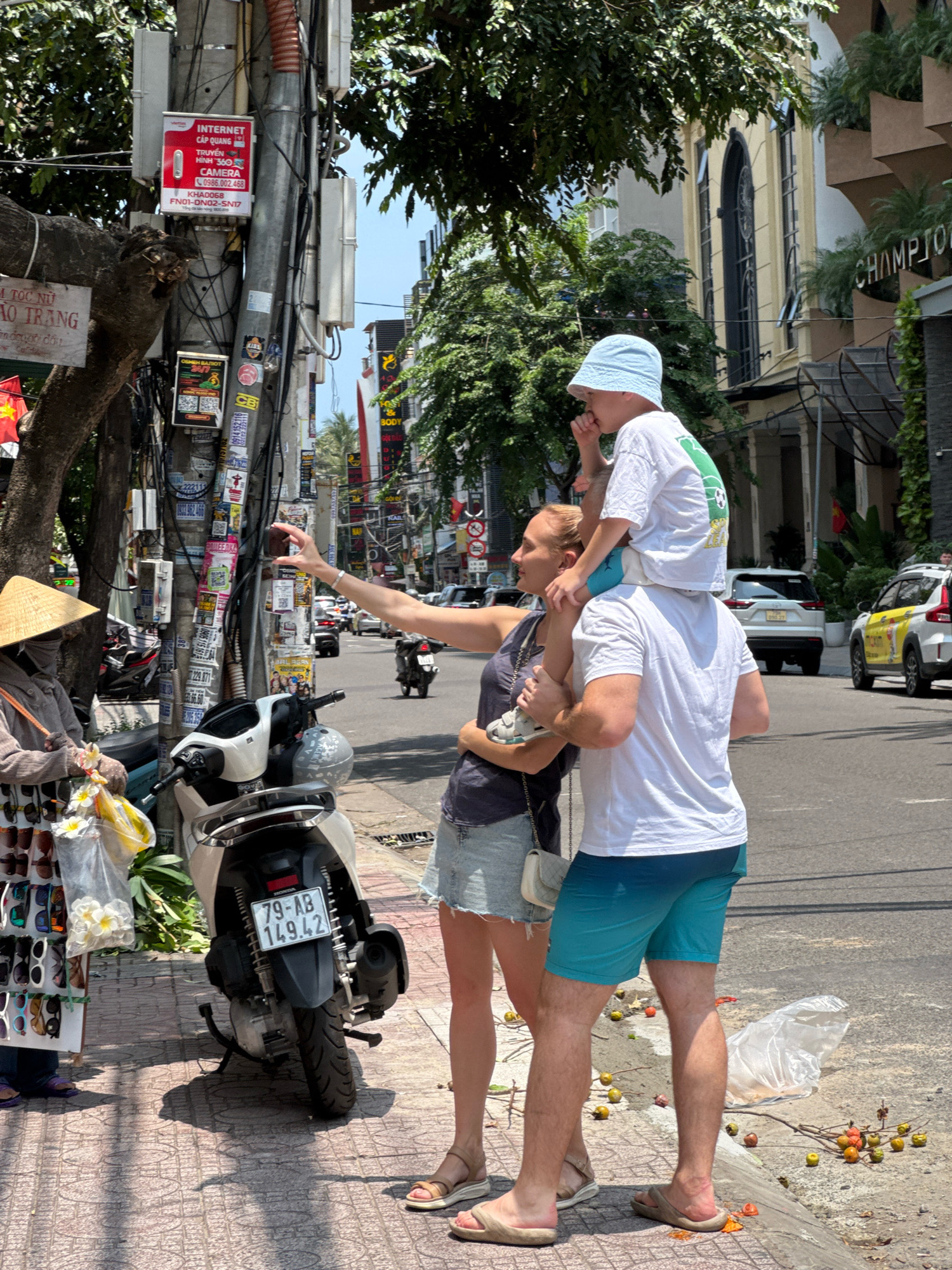 In Nha Trang, Vietnam, a family is captured on a city street. A young child, wearing a light blue bucket hat, sits atop the shoulders of an adult male who is facing away from the camera, wearing a white t-shirt and blue ombre shorts. Beside them, an adult female, with blonde hair pulled back, faces the camera and is reaching out with her left hand, holding a smartphone, as if taking a photo or video. She wears a dark tank top and a denim skirt. Next to the family, a vendor wearing a conical hat and face mask stands behind a display of sunglasses and flowers. A white scooter is parked to the right of the vendor and in front of the family. The street is paved with tiles, and a concrete curb is visible. Scattered on the curb are fallen fruits. The background shows a bustling city street with buildings, trees, other vehicles, and utility poles adorned with numerous signs and wires. The scene is bathed in bright sunlight, indicating a daytime setting.