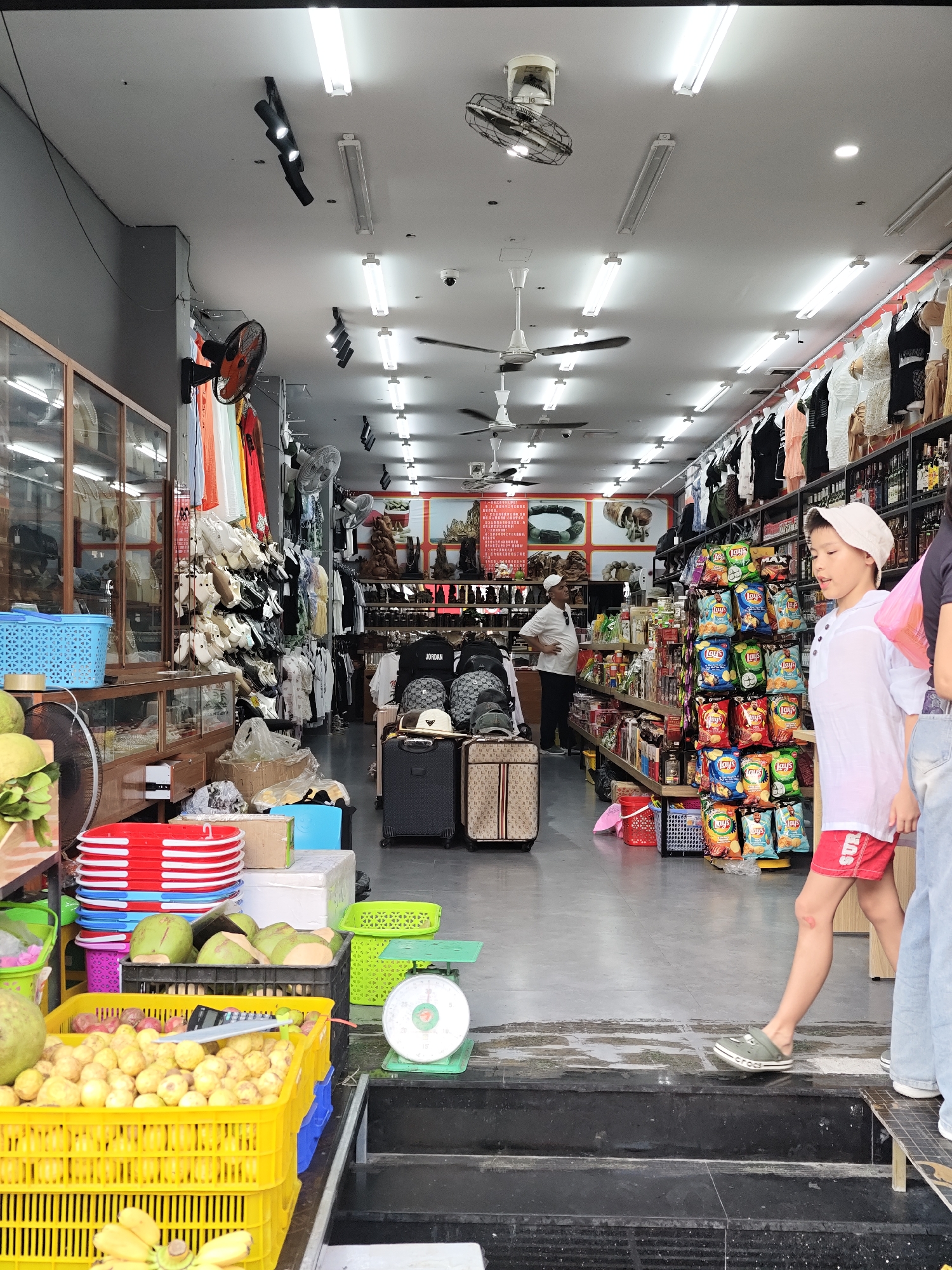 This is a medium shot, eye-level photograph of a store in Nha Trang Ward, Vietnam, that sells various goods including clothing, food, and other merchandise. In the foreground, there are stacks of yellow and blue plastic crates filled with fruits, red and blue stacked plates, and a scale. A young boy wearing a white shirt and red shorts is walking away from the camera, looking to his right. To the right of the boy, a person wearing blue jeans is visible. In the midground, there are aisles of shelves stocked with bagged snacks, clothing on hangers, and other miscellaneous items. A man in a white shirt and dark pants is standing near a shelf, facing away from the camera. In the background, the store extends further, with more shelves and merchandise visible. The ceiling of the store is equipped with fluorescent lights and ceiling fans. The overall impression is of a busy retail environment.