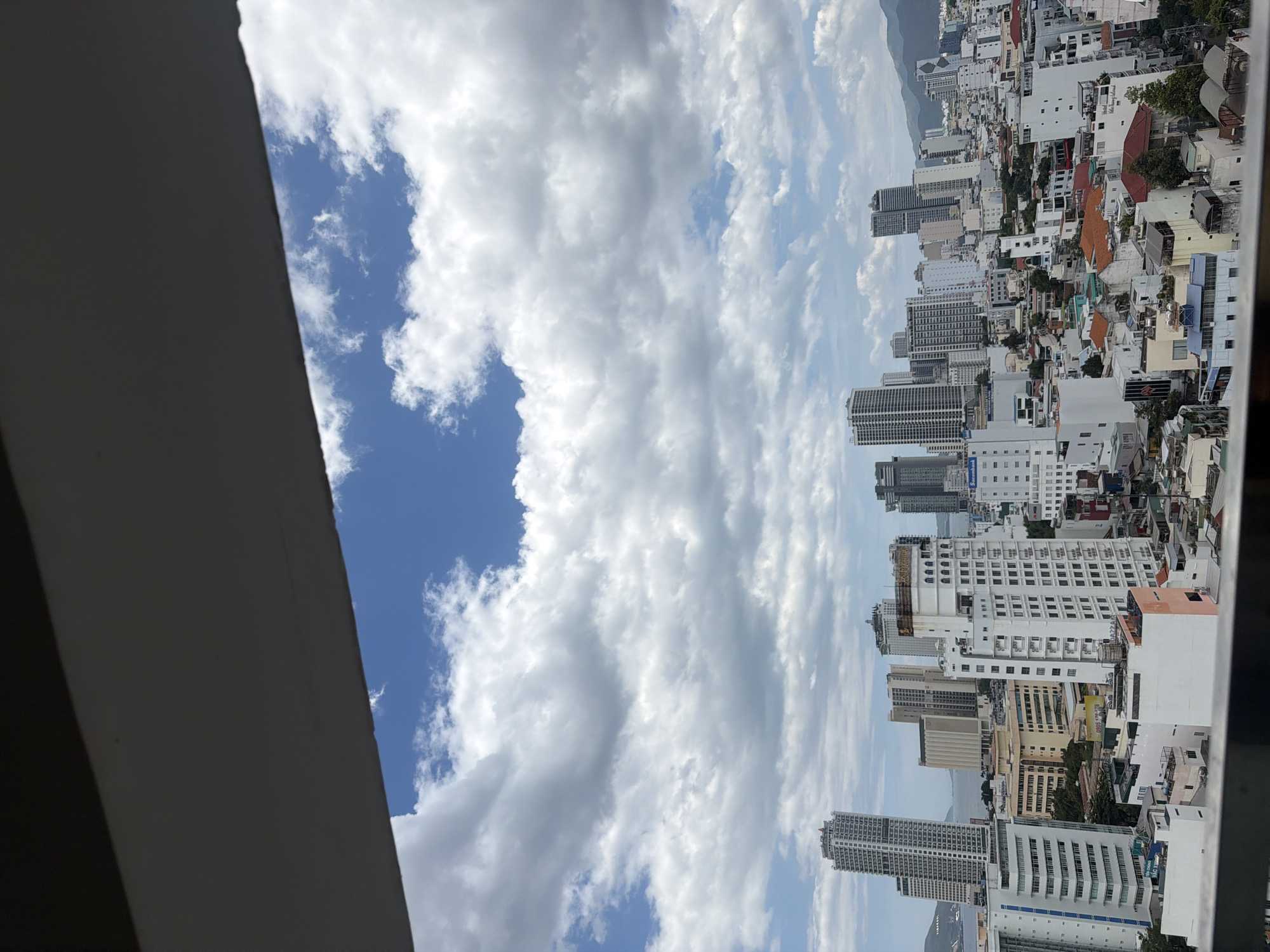 The image captures a panoramic view of a dense urban cityscape in Nha Trang, Vietnam, under a sky filled with dramatic white cumulus clouds against a bright blue backdrop. The city is characterized by a multitude of high-rise buildings, ranging in height and architectural style, dominating the landscape. Among these are several prominent skyscrapers, some with sleek, modern facades of glass and steel, and others with more traditional, textured exteriors. In the foreground, a dark, angled structure, likely a balcony railing or overhang, partially obscures the view.
The mid-ground and background reveal a tapestry of lower-rise buildings, many with terracotta or similar colored rooftops, interspersed with trees and greenery, suggesting a mixed-use urban environment with residential areas and commercial spaces. A body of water, possibly the sea, is visible in the distance, reflecting the cloudy sky, and rolling hills or mountains form the backdrop to the city. The overall impression is one of a vibrant, bustling coastal city with a high density of construction. No people are discernible in the image.