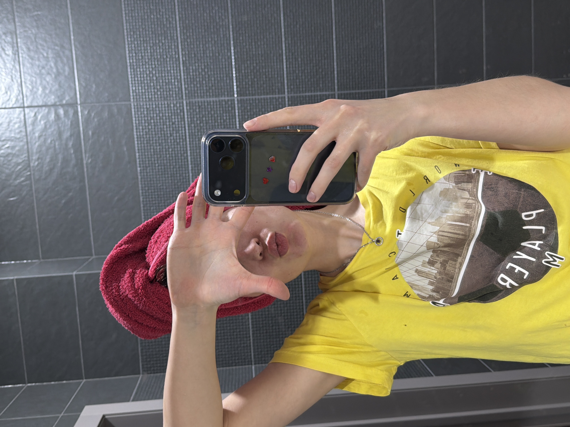 A person wearing a red towel on their head and a yellow t-shirt with a circular graphic is taking a selfie in front of a tiled wall. The person is holding a black smartphone with a clear case decorated with small heart-shaped stickers. The setting appears to be a bathroom. There is no information available to determine the specific location in Chaikovsky, Russia.