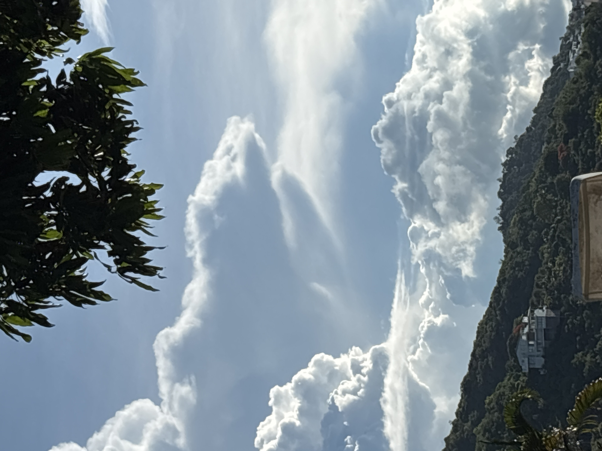 An outdoor medium shot shows a cloudy sky over a lush green hillside. The sky is a light blue, dotted with various white and gray clouds, some of which are large and billowy, while others are wispy and thin. In the upper portion of the frame, a cluster of dark green leaves from a tree hangs into view. The hillside is covered in dense green foliage, with a few trees and bushes visible. A modern white house with a reddish roof is perched on the side of the hill, its multiple levels and balconies clearly discernible. The overall scene evokes a sense of natural beauty and tranquility.