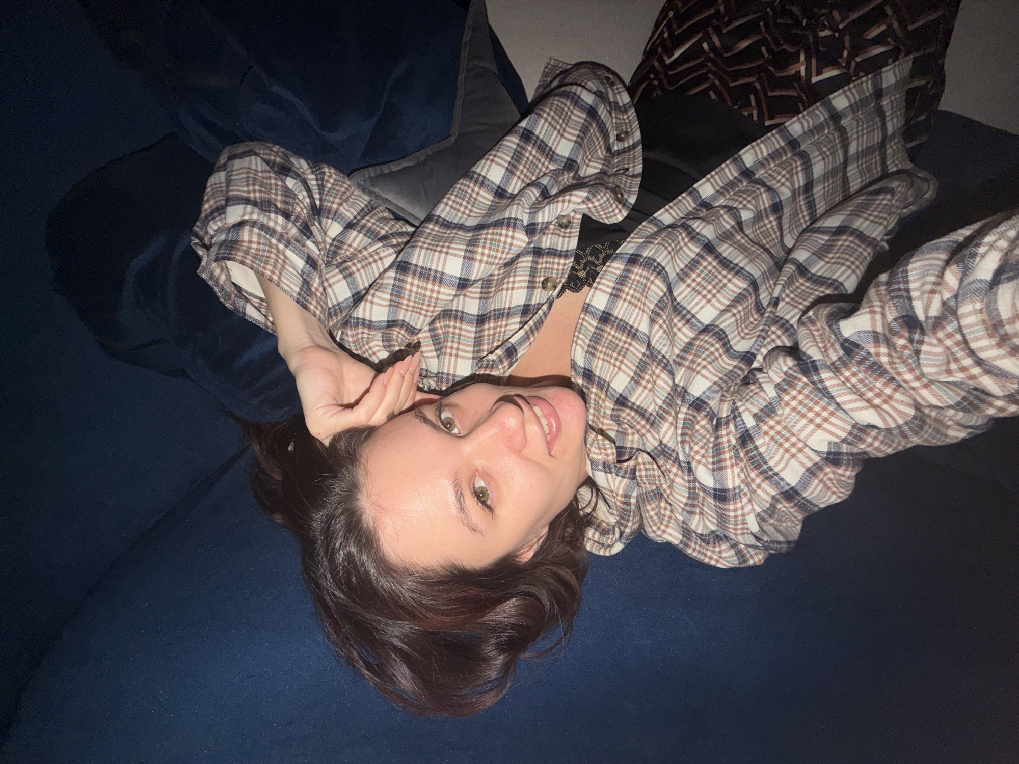 A woman is lying on a blue couch, wearing an open, plaid, button-down shirt. The photo is taken from above, with her head tilted toward the camera. Her hair is dark, and she is smiling, with her right hand near her forehead. A pillow is visible behind her head, and another patterned object is slightly visible in the upper right corner. The location is not discernible.