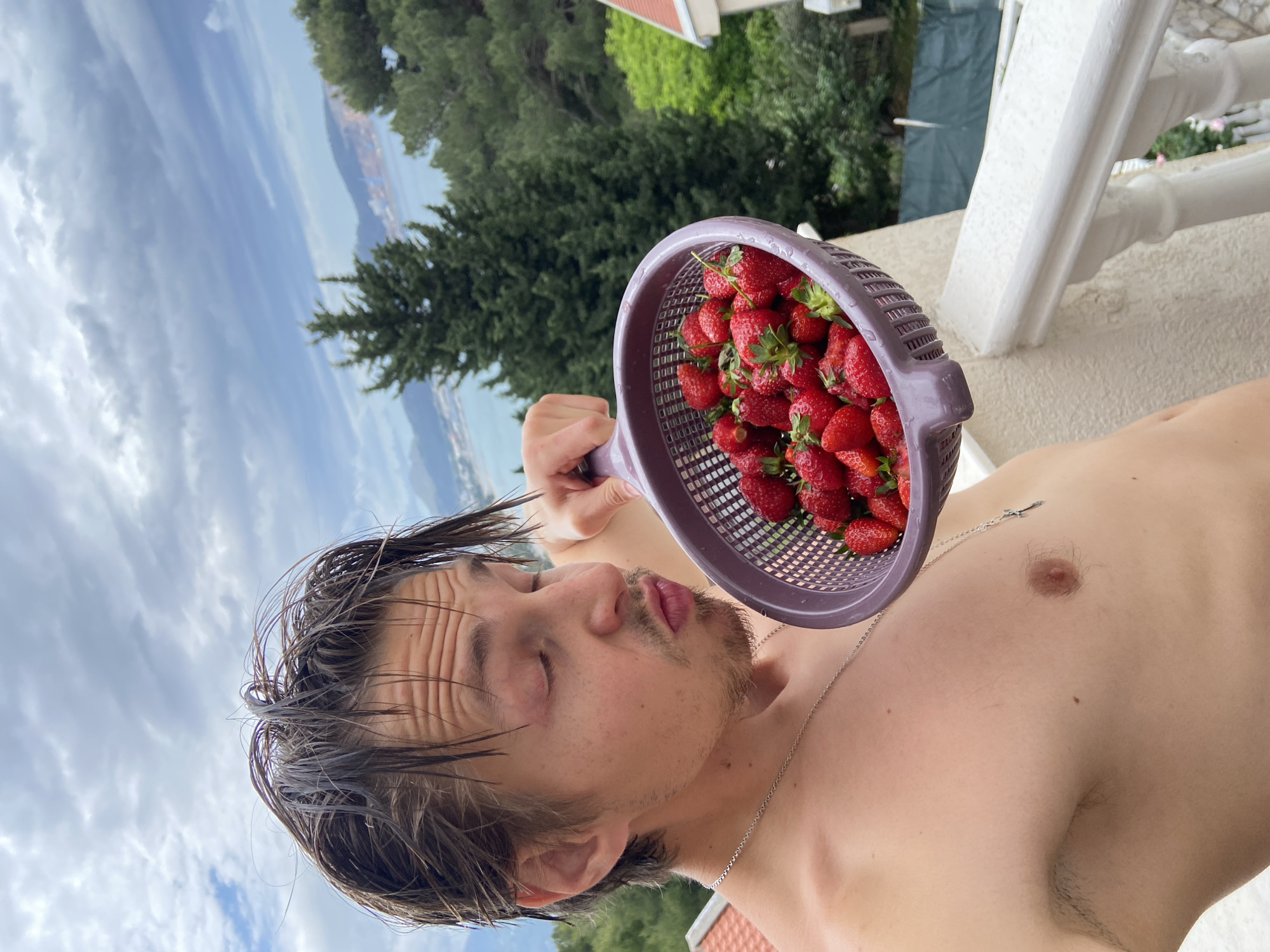 A shirtless man is holding a purple colander filled with strawberries near his face. He is facing left, with his eyes closed and his lips pursed. The background shows a cloudy sky, greenery, a body of water, and a glimpse of a building with a red roof. The scene is at an outdoor location, seemingly on a balcony with white railings. The location is Šušanj, Montenegro.