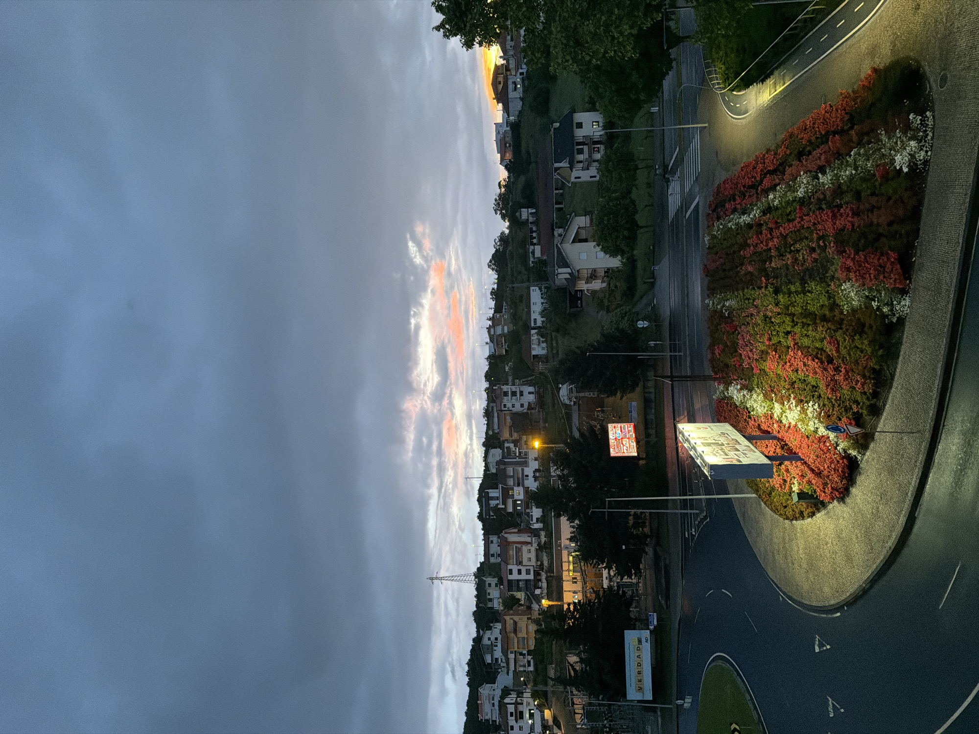 This image depicts a roundabout with a flower bed in Bragança, Portugal, at dusk. The sky is mostly overcast with hints of pink light. The flower bed has red, white, and green flowers. The roundabout is lit by streetlights. Residential buildings are on a hill in the background with a communications tower visible. There are commercial signs near the roundabout.