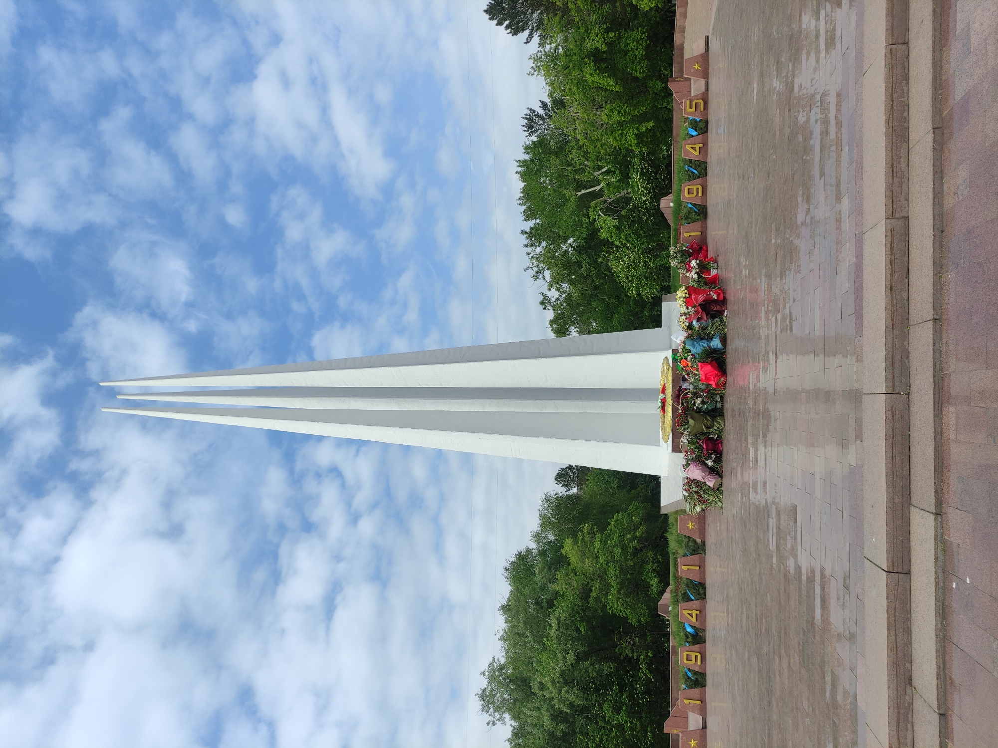 The image depicts a monument in Pavlodar, Kazakhstan, dedicated to the victory in World War II. The monument features three white, upward-pointing pillars against a blue sky with scattered clouds. At the base of the pillars are wreaths of flowers. To the right of the monument, the numbers 1941 and 1945 are displayed in gold lettering. The ground appears to be paved with red granite. The surrounding environment includes trees.