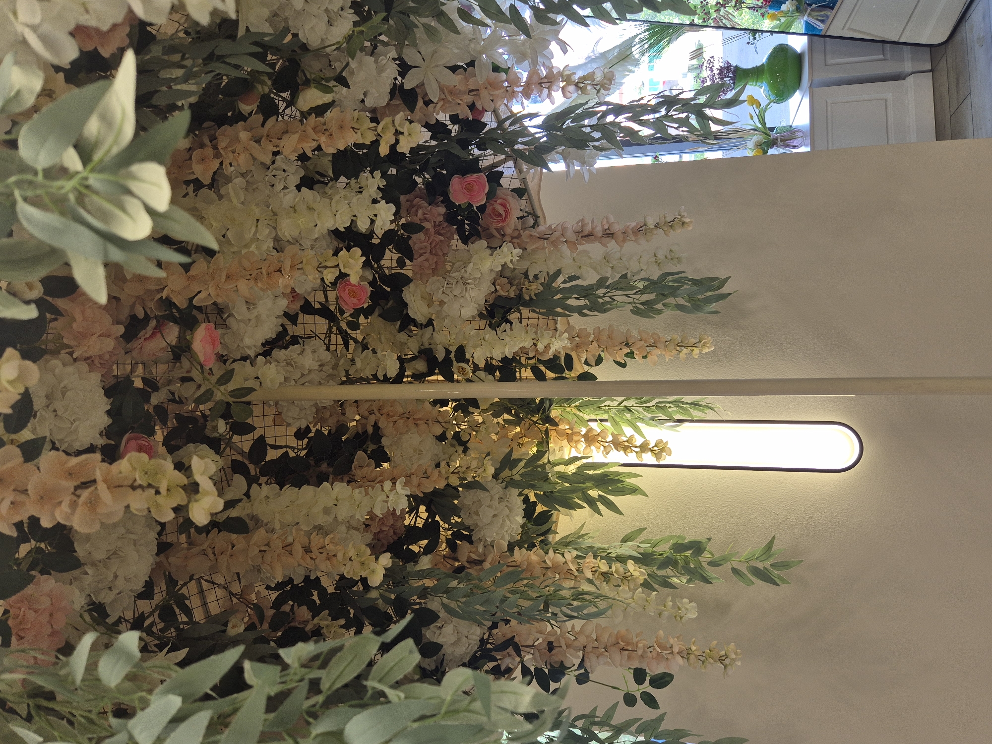 An indoor scene showcases a ceiling covered in a dense arrangement of artificial flowers and greenery. Cascading down from the ceiling are numerous floral strands in shades of white, cream, and pale peach, interspersed with realistic green foliage. A thin, vertical white pole is positioned centrally, supporting the floral display. To the right, a tall, elongated wall sconce emits a bright, diffused light. Behind the pole and to the right, a white wall is visible, with a window in the background revealing a glimpse of an outdoor area. On a white ledge beneath the window, several decorative vases are present, including a vibrant green vase holding a bouquet of flowers. The overall impression is one of opulent floral decoration. There is no specific indication of the city of Kokshetau, Kazakhstan in the image itself.