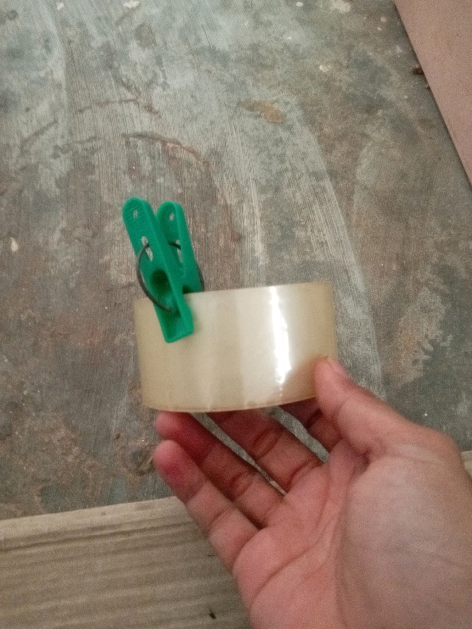A hand holds a roll of clear packing tape with two green clothespins clipped to its side. The tape roll is partially obscured by the hand from the bottom right corner of the frame. The background appears to be a textured, grey concrete surface. The scene is indoors, as suggested by the lighting and the presence of a wooden object in the bottom edge of the frame. The image is devoid of any identifiable landmarks or clues that would suggest a specific location within Batam, Indonesia.