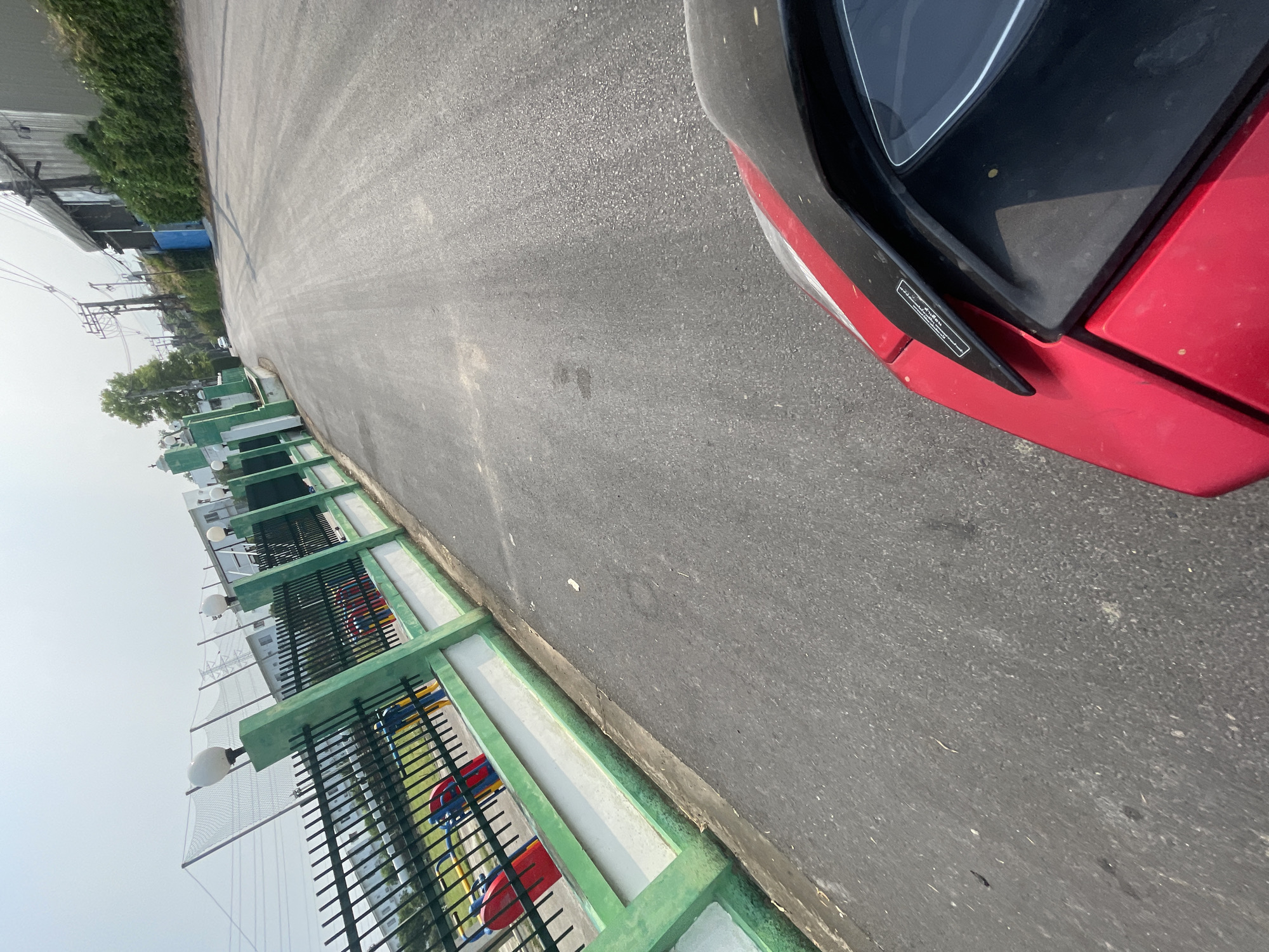 The image depicts a section of asphalt road, likely within the Nong Pa Khrang area of Thailand, given the prompt's instruction. A portion of a red and black vehicle is visible on the right side of the frame, occupying a significant amount of space. To the left of the road is a low fence with a green and white concrete base and black metal bars. Behind the fence, glimpses of what appears to be a playground are visible with red, blue, and yellow structures. Further in the background, possibly across the playground, are indistinct buildings, some partially obscured by trees. The sky appears overcast or hazy.