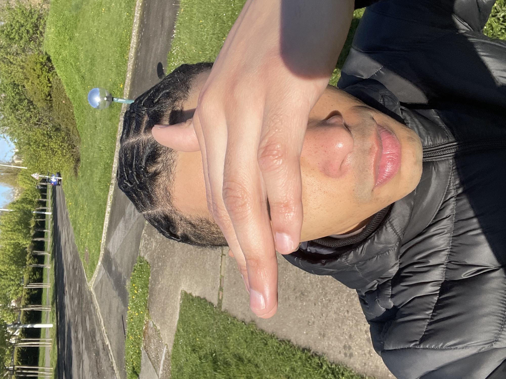 The image depicts a close-up, first-person view of an individual in an outdoor setting within Montigny-le-Bretonneux, France, likely on a clear, sunny day. The subject is a person of color, presenting as male, with dark, braided hair styled in cornrows with a visible central part. They are wearing a black quilted puffer jacket, visible from the upper chest to the shoulders. Their right hand is raised, palm partially facing the camera, with fingers slightly splayed, covering the upper half of their face (eyes and forehead) to shield from direct sunlight. The lower part of the face is visible, showing a nose with freckles/pores and pink lips. Strong sunlight illuminates the scene from the left, casting a distinct shadow from the hand onto the subject's face and jacket.
The background reveals a paved pathway or sidewalk in the immediate foreground, transitioning to green grassy areas with scattered small yellow flowers. Beyond this, an asphalt road or wider pathway curves through the midground. To the right, a silver spherical object on a light-colored pole stands within a grassy area. Further back, a gently sloping green mound with dense bushes is visible. In the distance, a line of uniformly spaced, slender trees with light trunks and green foliage forms a visual barrier, behind which a small blue car is partially visible near a triangular road sign. The overall environment suggests a well-maintained park or residential green space with pedestrian infrastructure.