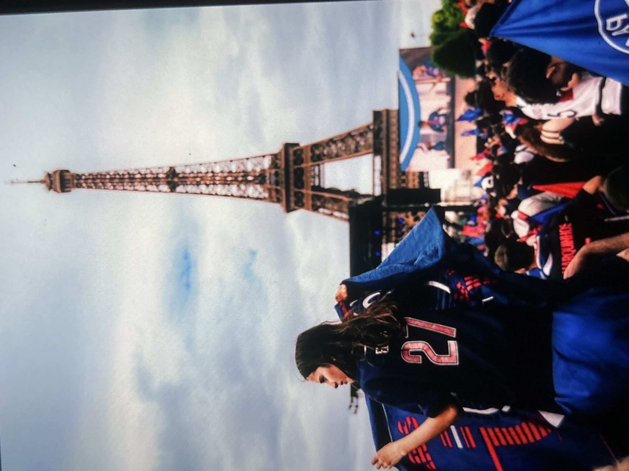 The image depicts a large outdoor gathering with the iconic Eiffel Tower dominating the background under an overcast sky. In the foreground, a person with dark hair, seen in profile, is wearing a blue sports jersey with the red number "27" clearly visible on the back. This individual is holding a blue flag, part of which features a white and blue logo, strongly resembling that of Paris Saint-Germain (PSG).
Behind the person in the foreground, a dense crowd of people is gathered, many of whom are also holding blue flags or banners, indicating a shared allegiance or celebration. A large screen is visible above and behind the crowd, displaying a scene or graphic, which also features blue and white elements, further suggesting a connection to a sporting event or team. The setting is clearly a public event, likely a viewing party or a celebration of a sports victory.
The time of day is daytime, evidenced by the natural, albeit soft, light filtering through the clouds. The general mood appears to be one of excitement or anticipation among the crowd.
It is important to note that while the provided location context is "Bania, Bulgaria," the unmistakable presence of the Eiffel Tower unequivocally places this scene in Paris, France.