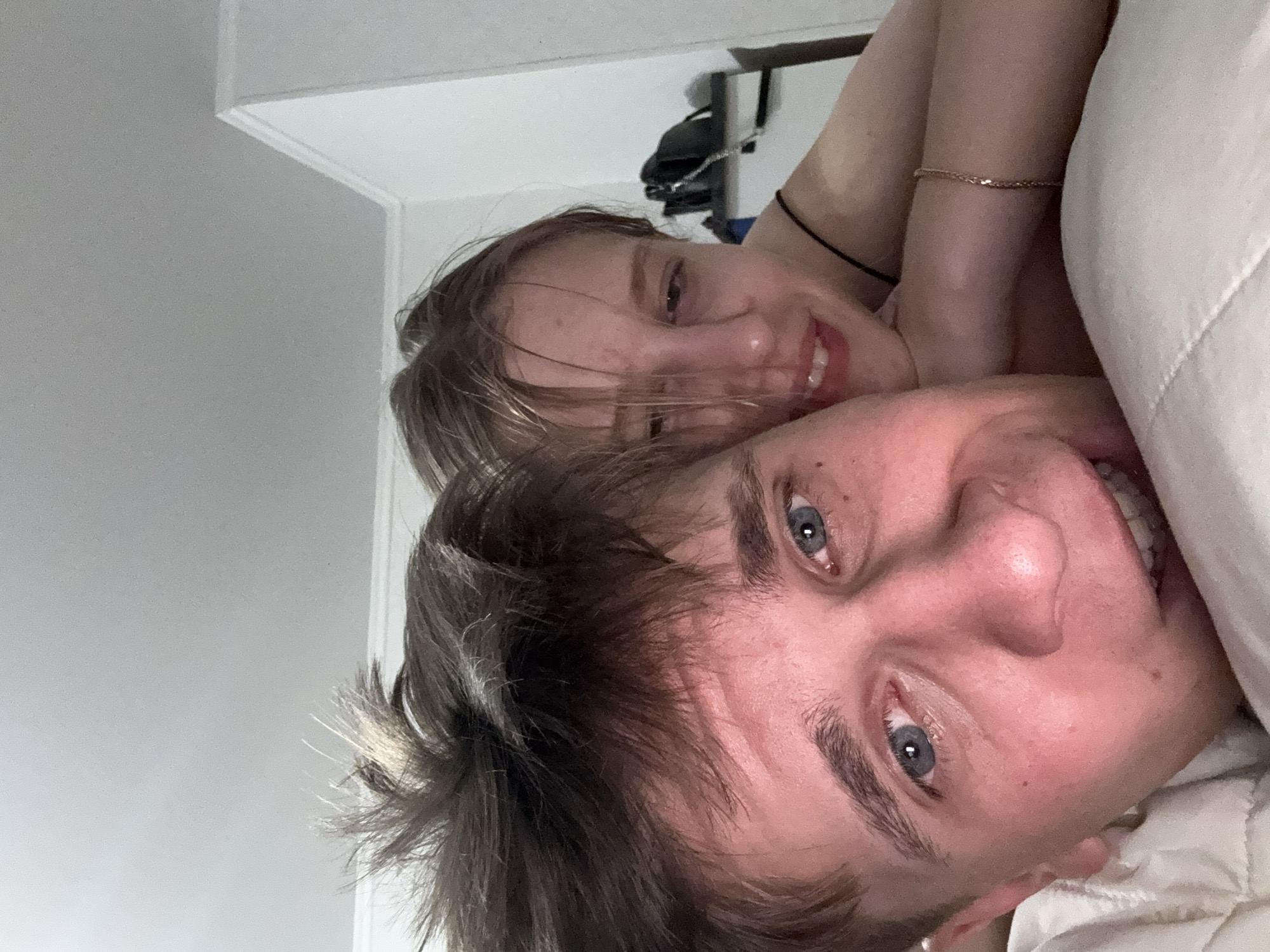 The image is a close-up selfie taken indoors, likely in a bedroom. Two individuals, a man and a woman, are visible. The man is in the foreground, looking directly at the camera with his eyes open and a slight smile. His hair is messy and light brown. The woman is behind him, her head resting on his shoulder. She is looking slightly away from the camera with a relaxed expression. Her hair is blonde. The background shows a white wall and a corner of the room, suggesting an indoor setting. There is a shelf with some items on it behind the woman. The lighting appears to be dim.