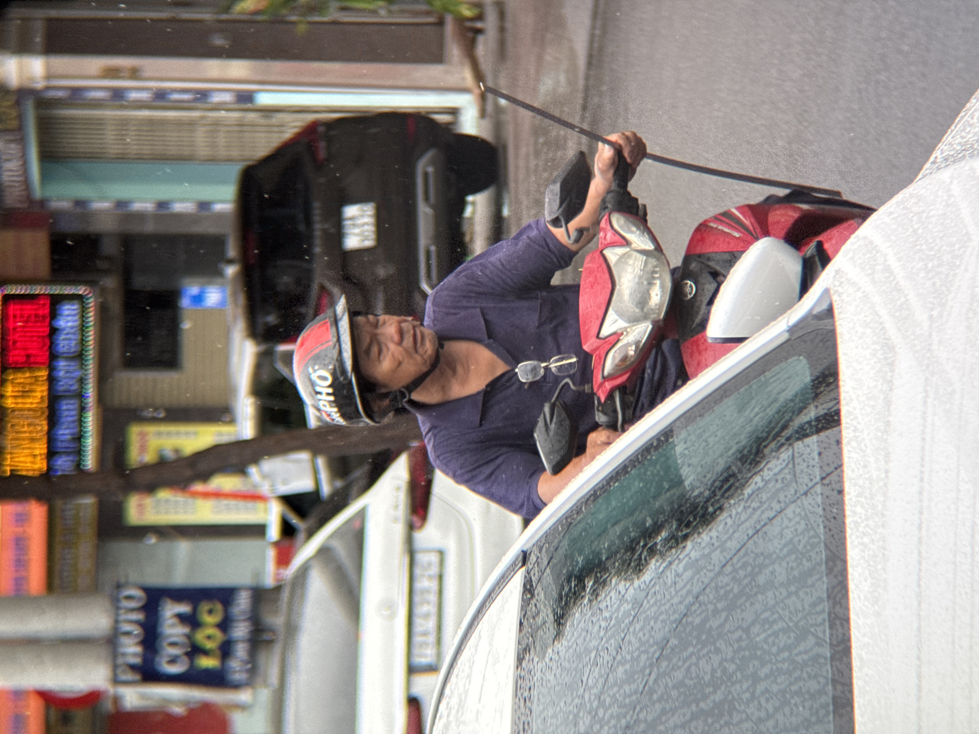 A man is riding a red and black motorbike on a street in Nha Trang, Vietnam. He is wearing a helmet with red and black accents, a dark blue polo shirt, and glasses. He is holding a long, thin black object, possibly an antenna or a flag pole, in his right hand. The motorbike has a prominent headlight and mirrors. In the background, there are other vehicles, including a white car and a dark SUV. Signs with Vietnamese text are visible on buildings. The scene appears to be wet, suggesting it might be raining, with water droplets visible on the windshield of a white car in the foreground and on the surfaces of the road. The man is looking forward as he rides.