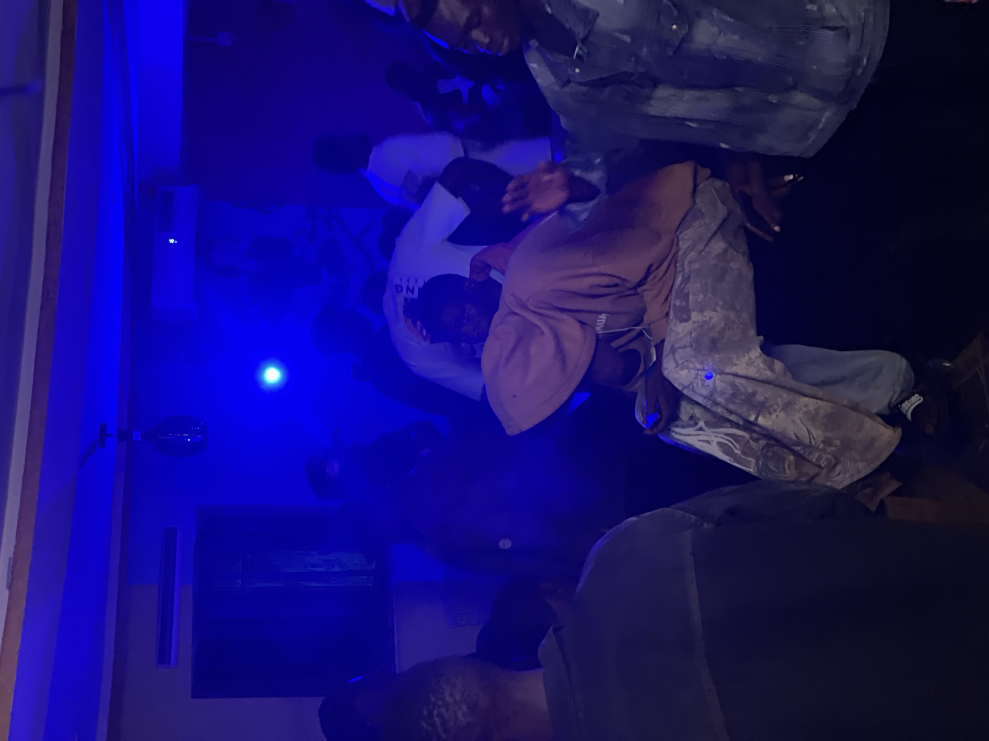 The image shows a dimly lit indoor space, possibly a club or lounge, with blue lighting casting an ethereal glow. Several individuals are present, engaged in various activities. In the foreground, a person wearing a light brown hoodie and patterned pants is seated, with their hand resting on their lap. To their right, another person in a denim jacket is standing, facing away from the camera, with their hand raised. In the background, more people are visible, some appearing to be dancing or socializing. The environment is characterized by dark walls and subtle architectural details, suggesting an urban setting in Durumi, Nigeria, perhaps a local entertainment venue. The overall mood is relaxed and casual, with a focus on social interaction and enjoyment.