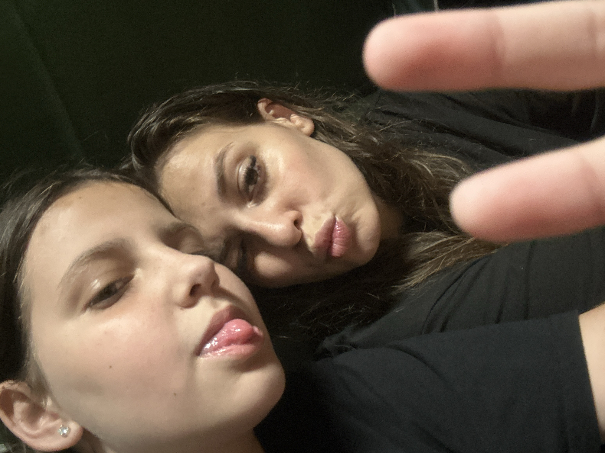 Two young women are captured in a close-up selfie. The woman in the foreground, on the left, has her head tilted, with her right ear visible and adorned with a diamond stud earring. Her eyes are partially closed, and her tongue is playfully extended. The woman behind her, on the right, is also leaning in, with her lips pursed as if for a kiss. Her eyes are also partially closed, and she has noticeable eyelashes. Both are wearing black shirts. The background is dark and indistinct. The image appears to have been taken indoors, possibly in a residential setting in Govedare, Bulgaria.