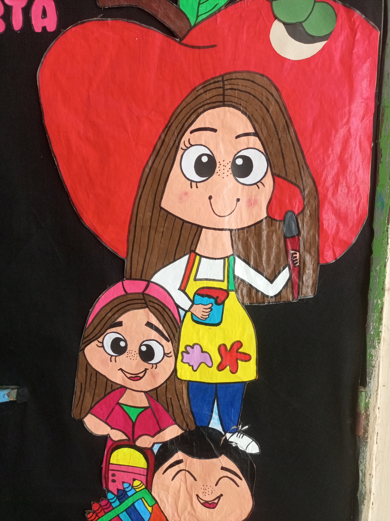 This image depicts a colorful, hand-drawn mural on a black background, likely displayed on a wall in a public space in Santa Ana, Venezuela. The mural features stylized cartoon characters, predominantly children, with exaggerated features like large eyes and simple, smiling expressions.
In the foreground, a young girl is depicted wearing a pink headband, a pink jacket over a green shirt, and holding a red and yellow backpack filled with colorful crayons. To her right, and slightly behind her, another child's smiling face is visible, with eyes closed in happiness and a smattering of freckles.
Above them, a taller figure, presumably an adult or older child, stands with a bright yellow apron over a white shirt and blue pants. This figure holds a blue cup and a paintbrush in their hands, suggesting an artistic theme, perhaps related to a school or community art project. Behind this central figure is a large, red, heart-shaped object, resembling an apple, with green leaf accents. The word "TA" is visible in pink lettering on the upper left side of the mural, possibly part of a larger word or phrase.
The overall style is playful and childlike, with bold outlines and vibrant colors. The mural appears to be made from cut-out paper or fabric, adhered to a dark, possibly felt or canvas-like surface. The context suggests a cheerful and educational environment, typical of a classroom, community center, or a local event in Santa Ana, Venezuela. The presence of a slightly worn, painted wall edge on the right side of the image indicates it's mounted on a building or partition.