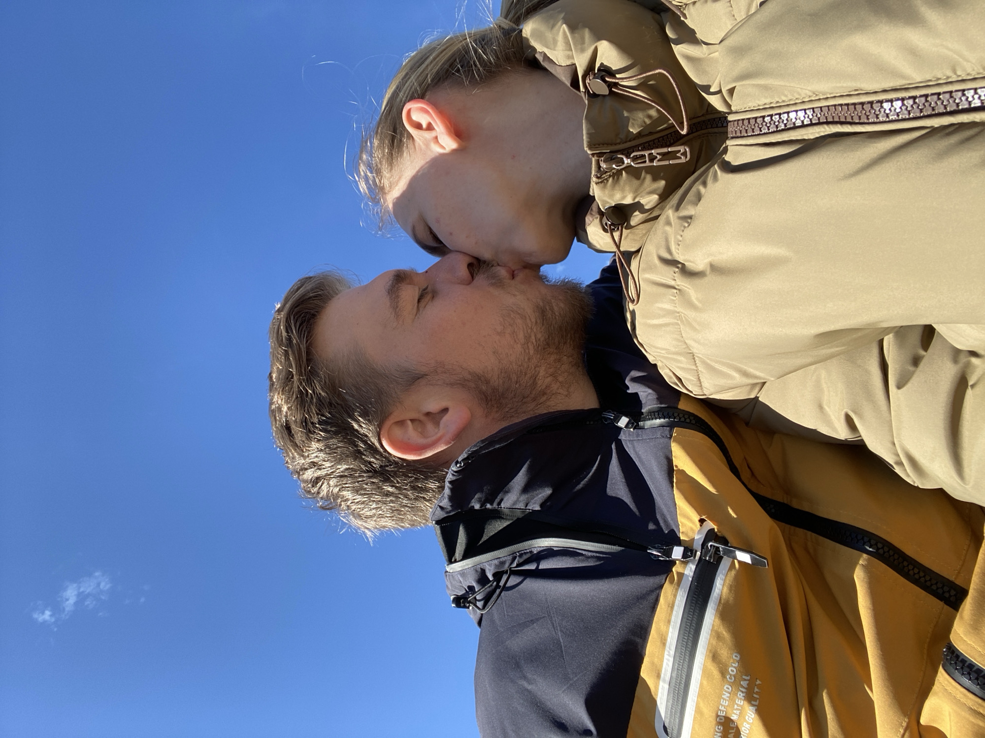 A man and woman are kissing against a clear blue sky. The man has short brown hair and a beard. He is wearing a blue and yellow jacket. The woman has blonde hair pulled back in a ponytail and is wearing a tan puffer jacket. The location is potentially Chaikovsky, Russia.