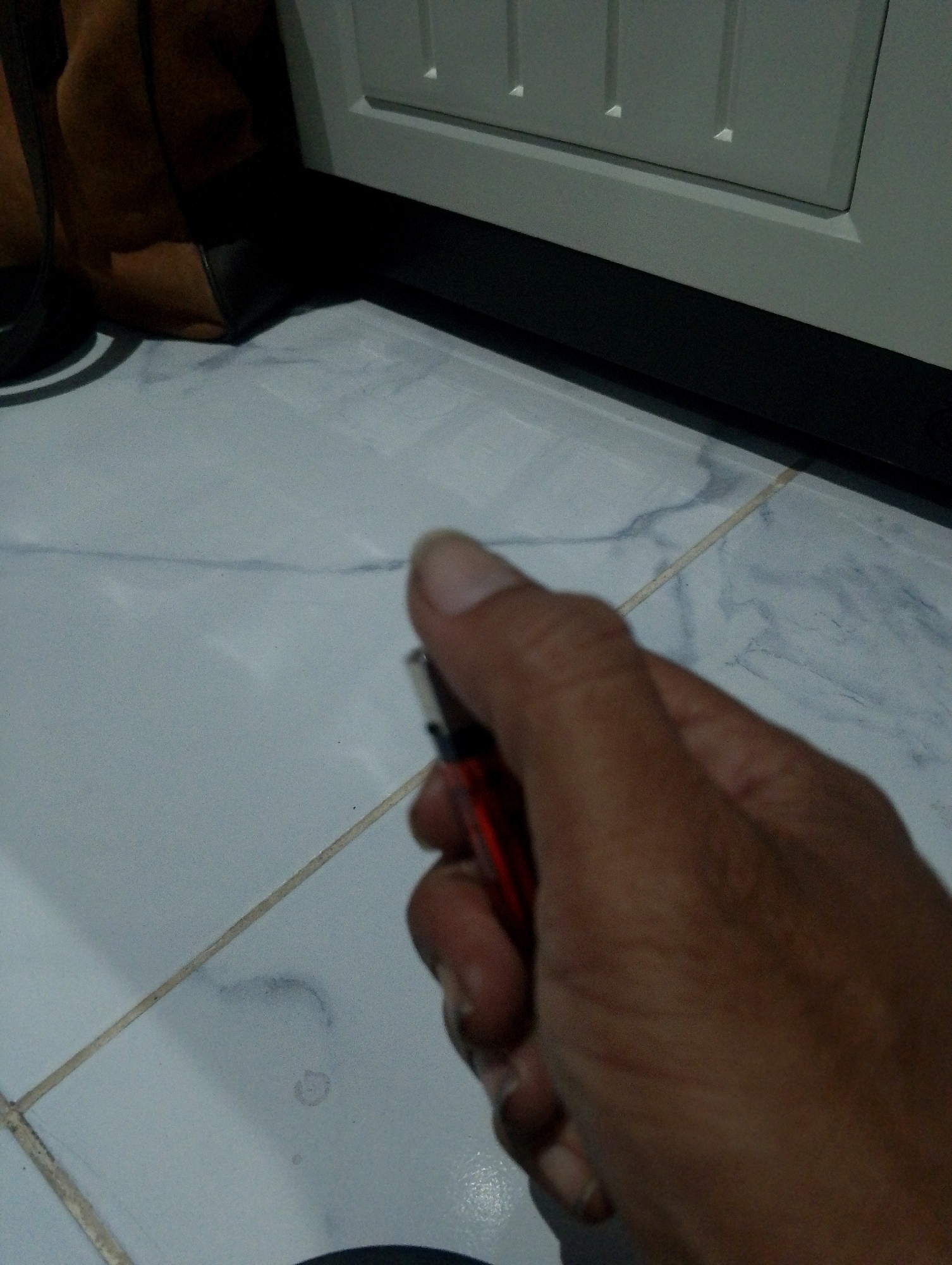 In the city of Sumbergirang, Indonesia, a close-up shot focuses on a person's right hand holding a red lighter. The hand, with visible veins and fingernails, grips the lighter firmly, suggesting an imminent action or a pause in one. The lighter, a common ignition tool, appears to be made of plastic and metal. The background reveals a tiled floor with faint grout lines, suggesting an indoor or covered outdoor setting. A dark, possibly leather, bag is partially visible on the left, and a portion of a light-colored cabinet or appliance occupies the upper right corner. The lighting is dim, casting soft shadows and highlighting the textures of the hand and the surrounding surfaces.