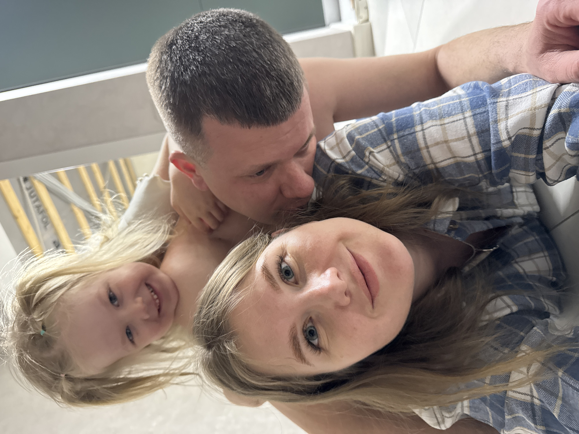 The image shows a selfie taken by a woman with light blonde hair, wearing a blue and white plaid shirt. She is smiling slightly at the camera. A man with short, graying hair is behind her, kissing her head. A young girl with blonde hair is also present, smiling at the camera. The background appears to be an indoor setting, possibly a bathroom, with a window or light fixture visible. It is not possible to determine the location in the city of Gornyi (Dvurechenskoe S/P), Russia from the given image.