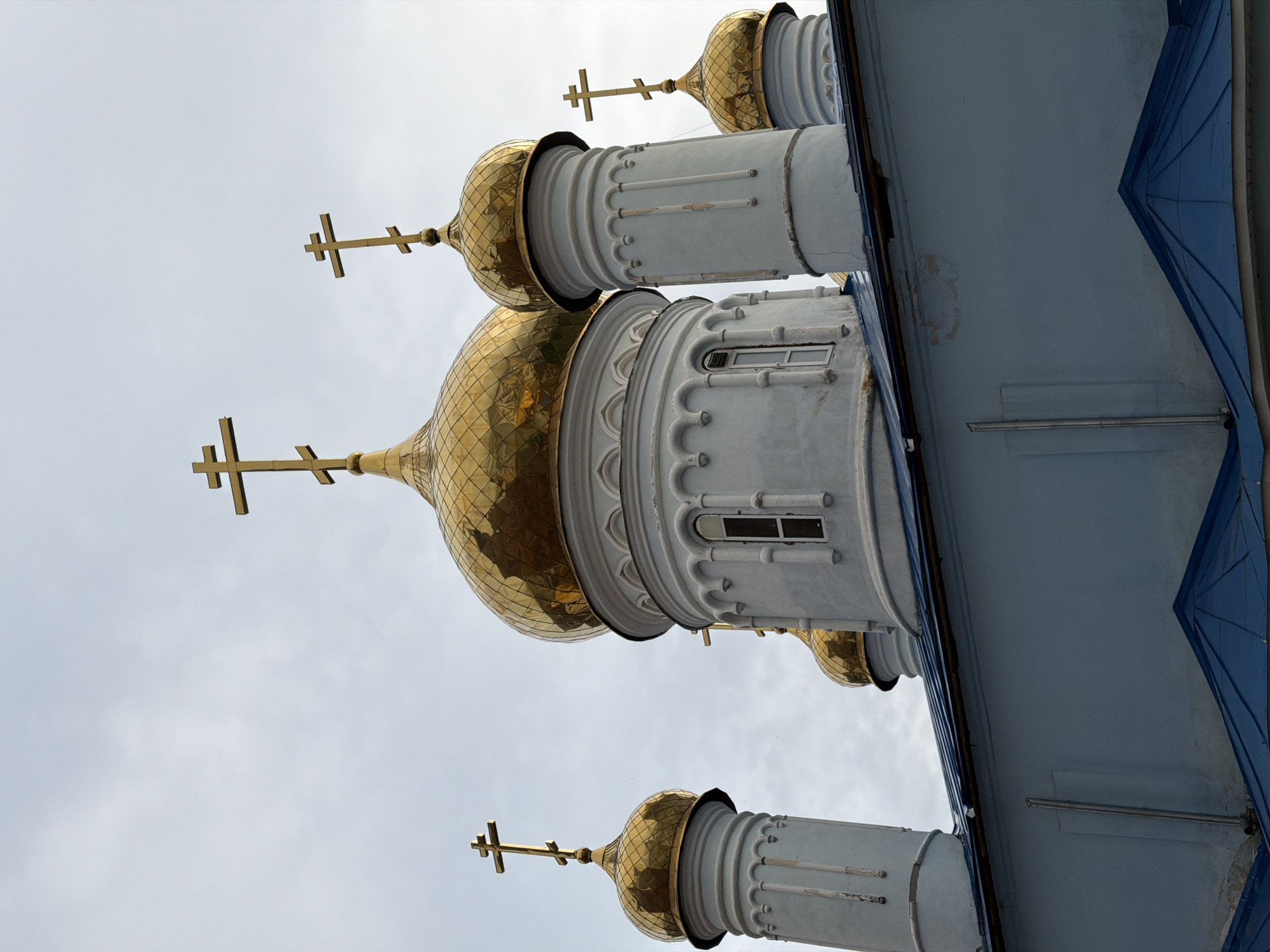 The image shows a low-angle view of the golden domes and crosses of the Annunciation Cathedral in Kazan, Russia. The domes are highly reflective, and some of the white cylindrical bases feature small windows. The sky is overcast. The side of the cathedral is visible, with blue roofing.