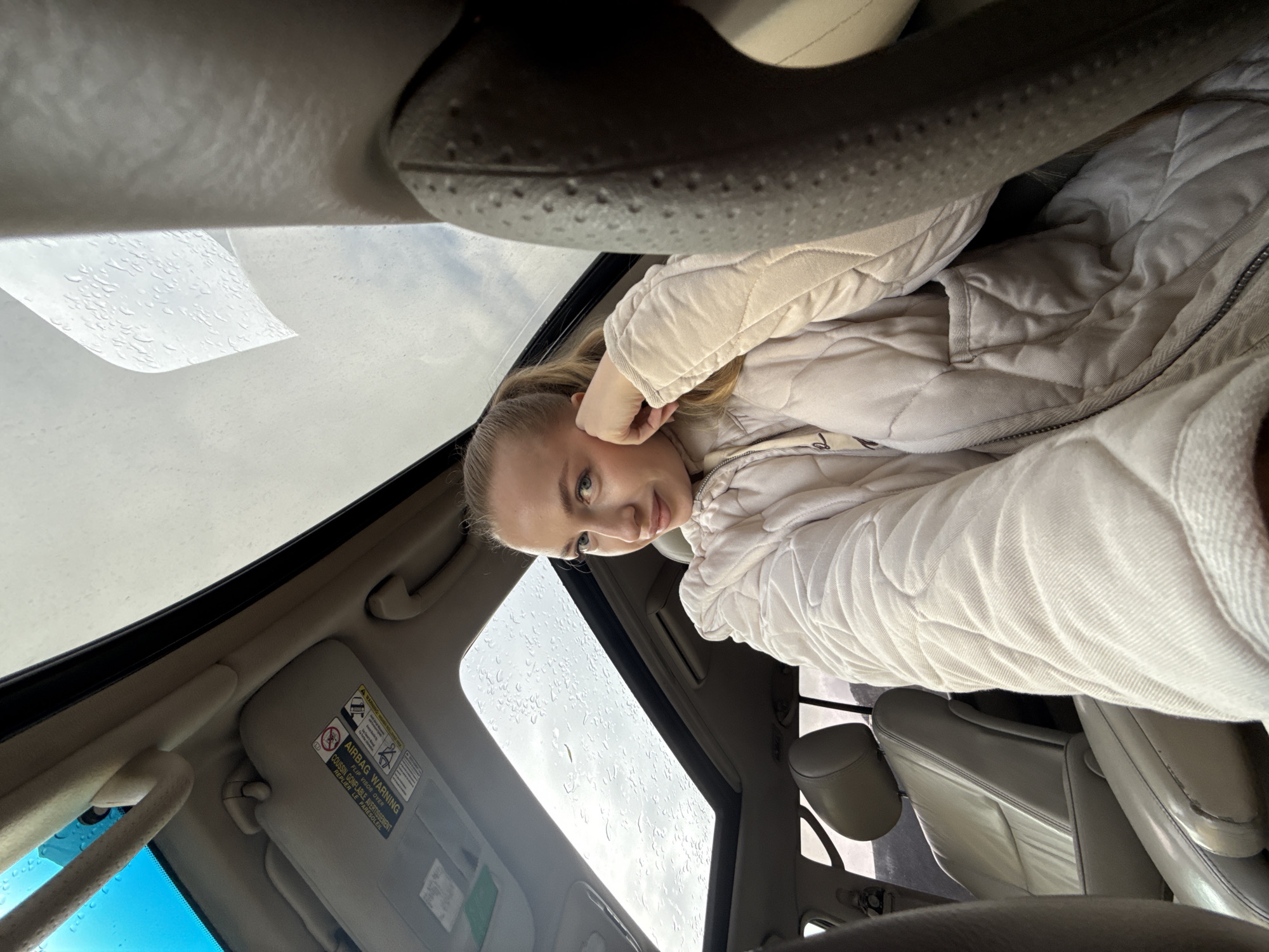 A woman is in a car with a sunroof. Rain is visible on the sunroof glass. The interior of the car is light gray or beige. The woman is wearing a light-colored quilted jacket and resting her head on her hand. A sun visor is visible. There is no indication of the location being Petropavlovsk-Kamchatsky, Russia, or any city.
