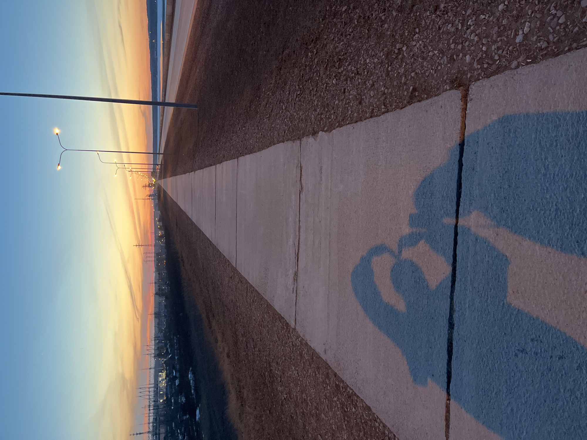 The image, taken in Chaikovsky, Russia, shows a sidewalk extending into the distance, flanked by a gravel area on one side and a view of the city illuminated by a sunset on the other. Streetlights line the sidewalk. The shadow of a person or persons making a heart shape with their hands is cast on the sidewalk in the foreground. The sky displays a gradient of colors from blue to orange. Buildings and power lines are visible in the distance.