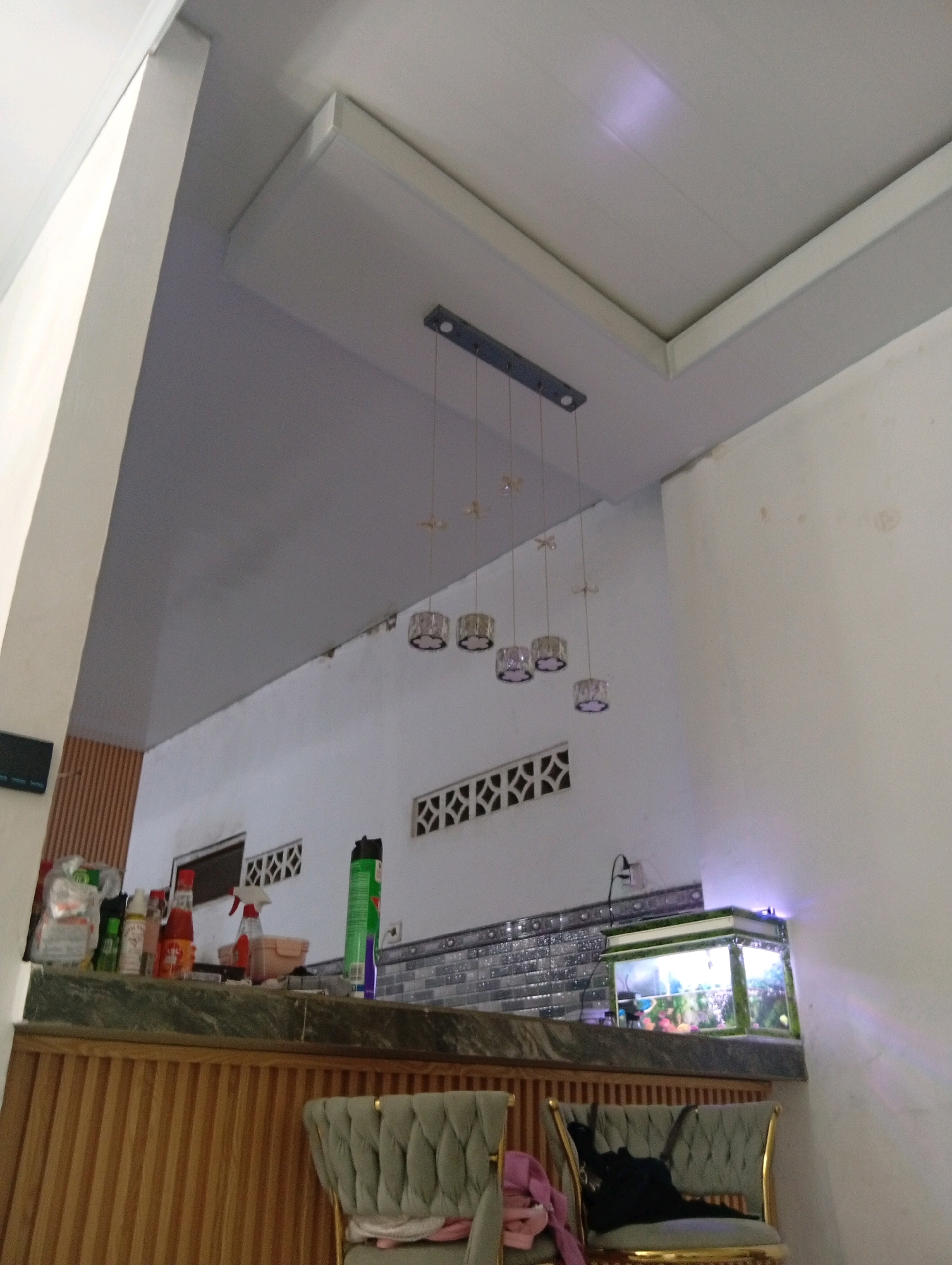 The image depicts an indoor scene, likely a dining area or kitchen, with a focus on a bar-like counter and overhead lighting. A multi-bulb pendant light fixture hangs from a recessed ceiling. Below the light, on a counter, are various items including bottles of condiments, cleaning supplies, and an aquarium filled with fish and aquatic plants. The counter itself appears to be made of dark marble and is supported by a wooden cabinet with vertical slatted detailing. Two upholstered chairs with a tufted back and gold trim are positioned in front of the counter. The walls are white, with some decorative ventilation grilles visible. The lighting suggests it is daytime or artificial light is on. The image is a static shot without any discernible human activity. The provided context places this scene in Samarinda, Indonesia.