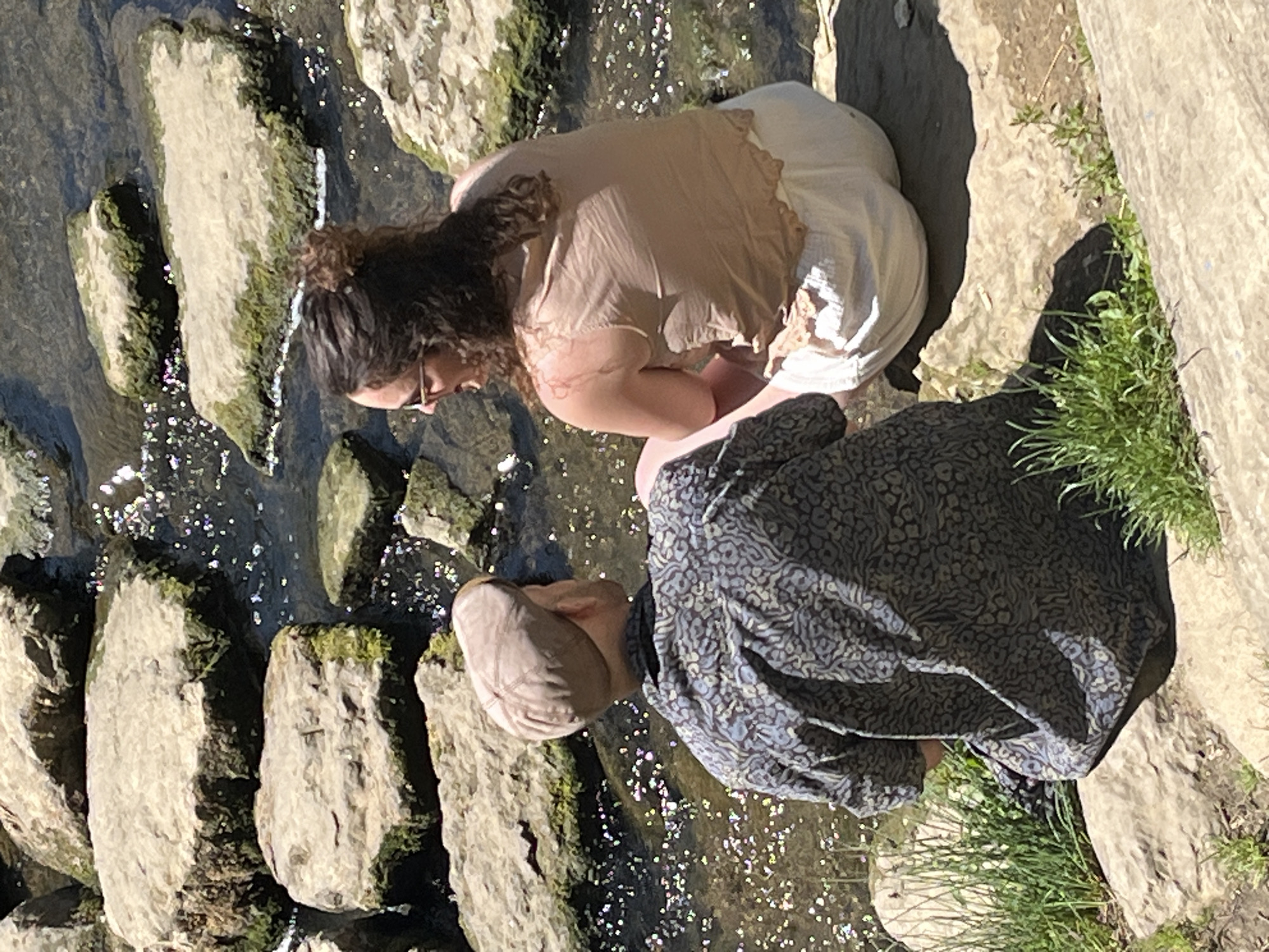 Two individuals are kneeling near a stream, seemingly engaged in an activity related to the water. The first individual is wearing a light-colored top and white pants, and has curly brown hair. The second individual is wearing a dark, patterned shirt and a light-colored cap. Both are positioned close to the water's edge, which is lined with rocks and grass. The stream itself is clear, with sunlight creating a sparkling effect on the water's surface. The environment includes rocks, grass, and flowing water. The image appears to be taken outdoors in natural light, likely during the day. The location appears to be the city of Luxembourg in Luxembourg.