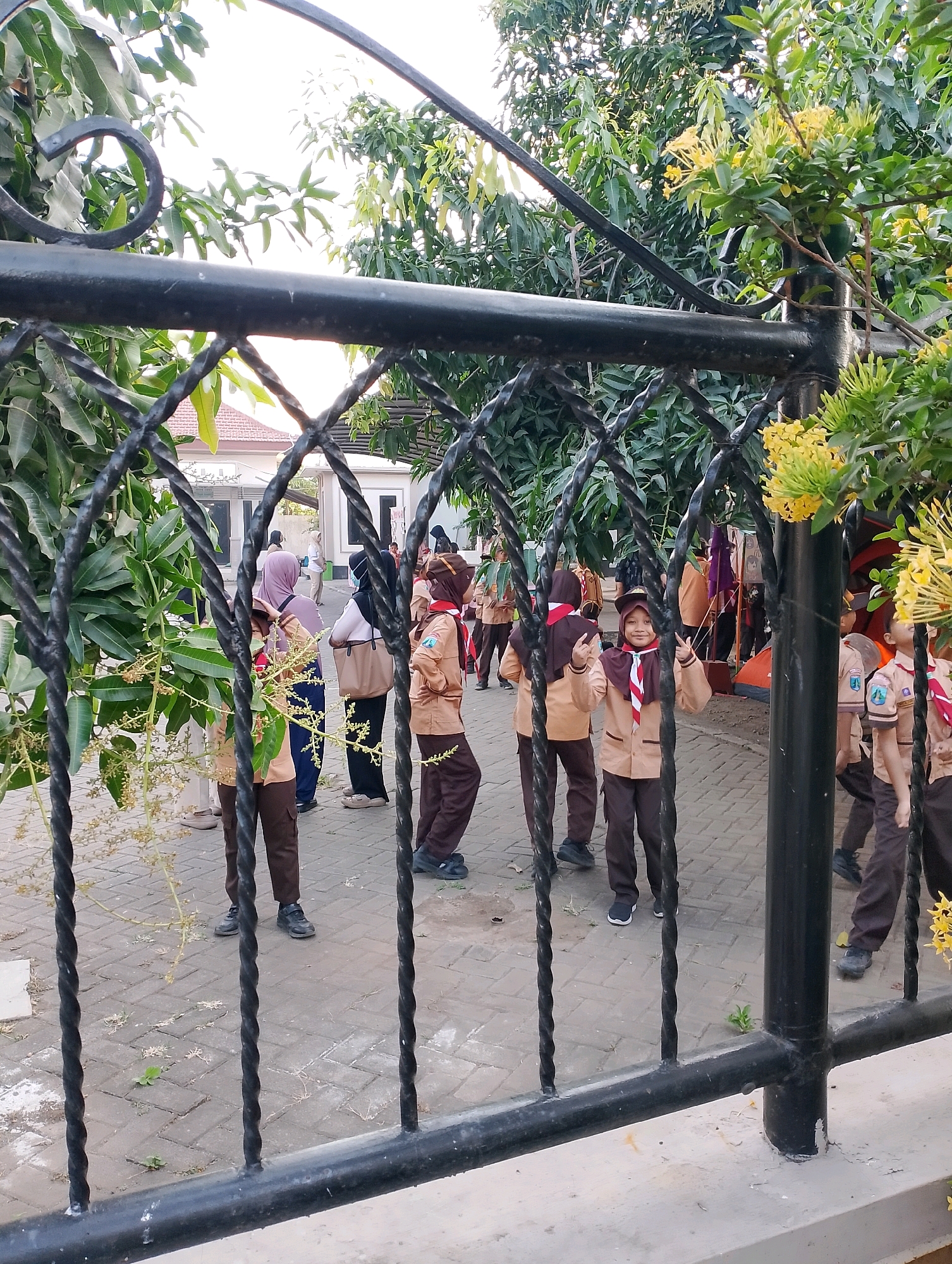 A group of Indonesian schoolchildren, likely from a Scout troop, are gathered outdoors behind a black metal fence. The children are wearing tan uniforms with brown pants and scarfs. Some are looking towards the camera, and one is making a peace sign with her fingers. The background shows trees, buildings, and other people, suggesting a school or community setting in Probolinggo, Indonesia. The fence is ornate, with twisted metal bars and a horizontal top rail. The ground is paved with rectangular stones.