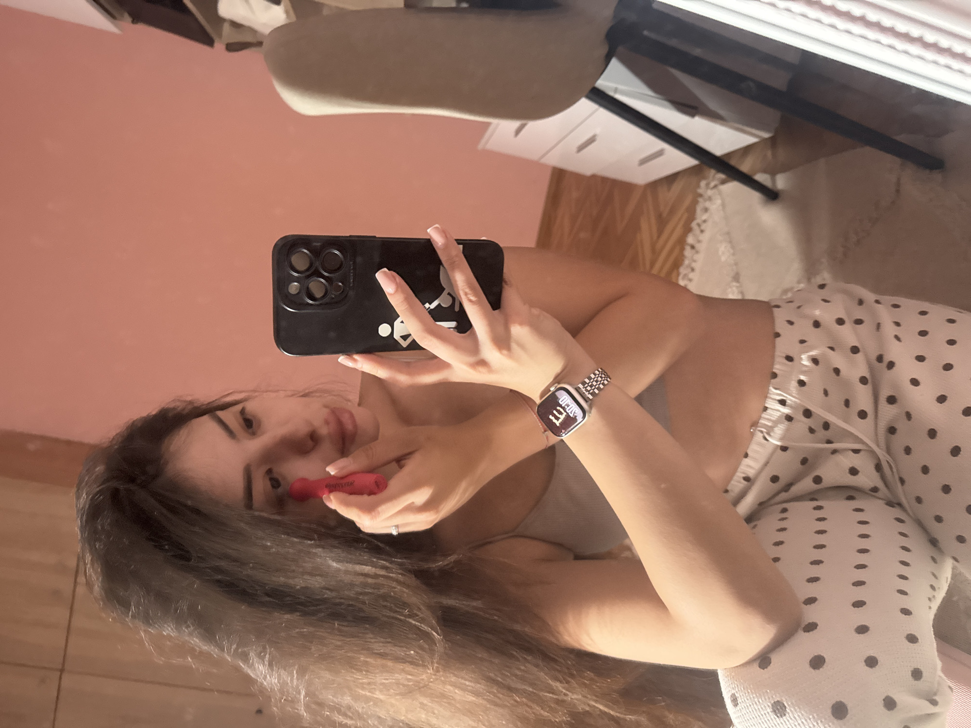 A young woman is depicted in a mirror selfie, oriented sideways across the frame, taken indoors in Plovdiv, Bulgaria. She is shown from the chest up, with long, dark hair.
The woman is taking a photo with a black smartphone, which has a triple-camera setup and a white stylized graphic on its case. With her other hand, she holds a red lipstick or lip gloss applicator near her lips, either applying makeup or posing. Her fingernails are well-manicured with light tips. She is wearing a light-colored top adorned with black polka dots.
On her left wrist, she wears a silver-toned metal link watch that displays the digital time "07:07", with faint text suggesting "THU 17" (Thursday 17th) and possibly "APPLE" on the watch face. The back of the phone also has subtle "APPLE" branding, and the word "Sephora" is faintly visible on the red applicator.
The reflected background features a light pink wall, a light-colored chair, white drawers, and the black legs of a table or desk. A patterned rug is also visible on the floor. Her expression is focused and calm.