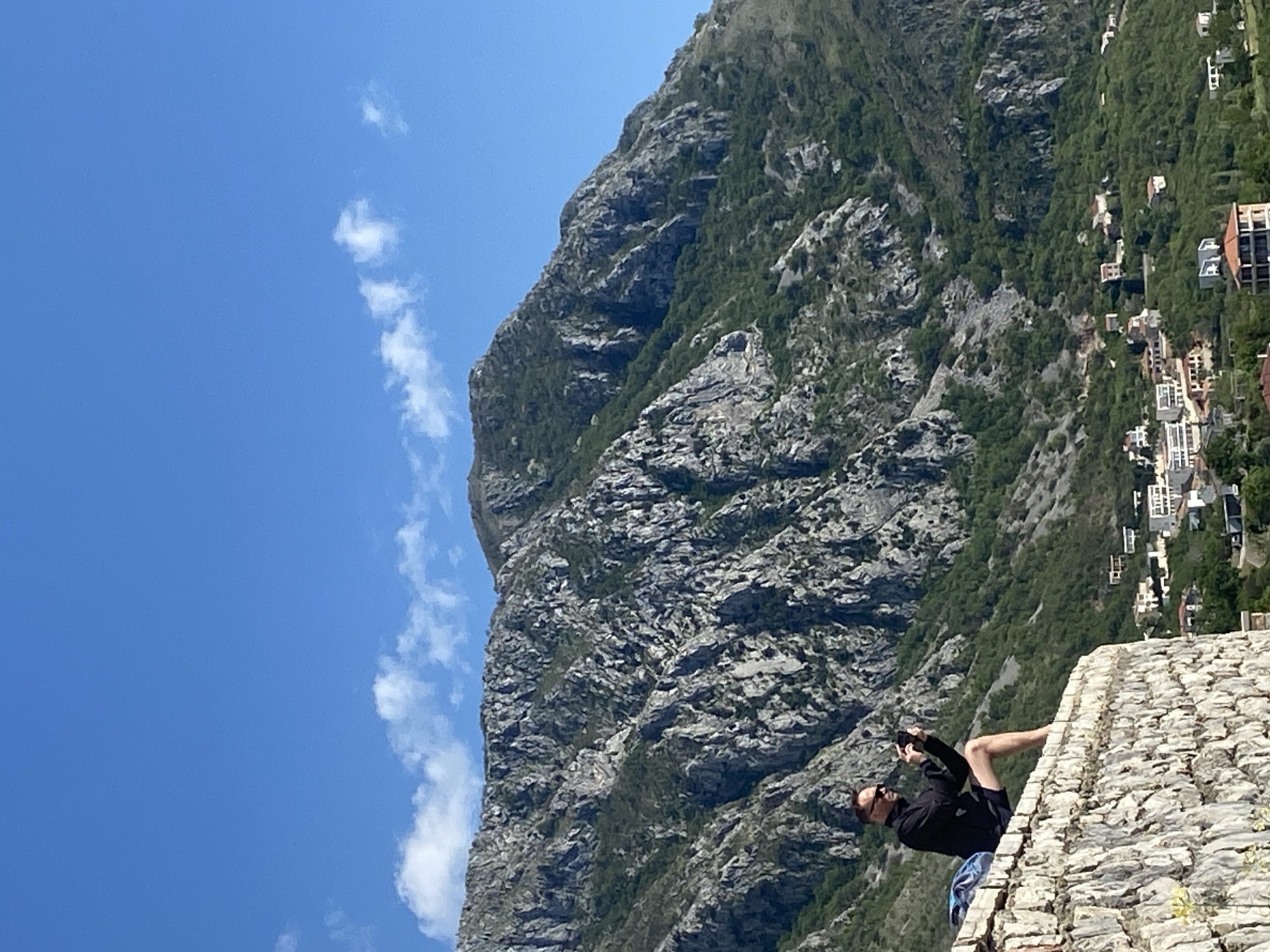 A full shot of a person taking a picture. The person is facing away from the camera, wearing a black long-sleeved shirt, sunglasses, and black shorts. The person is lying prone on a stone wall. The stone wall is made of irregular shapes and sizes of stone. In the background is a tall mountain covered in rocks and foliage. On the right side of the mountain is a small town with white buildings. The sky is blue with a few white clouds. The picture appears to have been taken in Sveti Stefan, Montenegro.