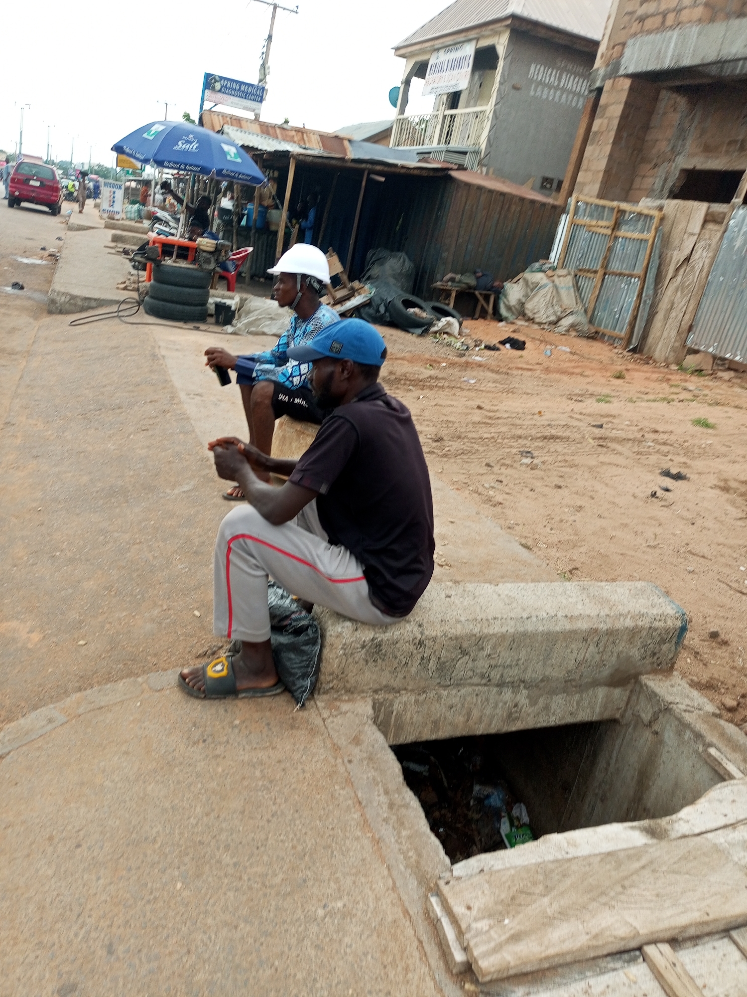The image depicts an outdoor scene in what appears to be a commercial area in Tempure, Nigeria. Two men are seated on a concrete ledge in the foreground, facing away from the camera and towards the street. The man on the left wears a white hard hat and a blue and white patterned shirt. The man on the right wears a blue cap and a dark t-shirt with light-colored pants. Both men appear to be holding objects, possibly phones, in their hands.
Behind the men, there is a street with a red vehicle visible in the distance. A blue umbrella with white lettering is set up, possibly for a stall. Several structures, appearing to be informal shops or buildings, are present. These structures are constructed with corrugated metal and wood, with some appearing weathered. A utility pole with wires is visible in the background, along with signage that includes text in English, such as "SPING MEDICAL DIAGNOSTIC CENTER". The ground is a mixture of dirt and concrete. A concrete drain opening is visible in the lower right of the image, containing debris. The overall environment suggests a busy, informal urban setting.
