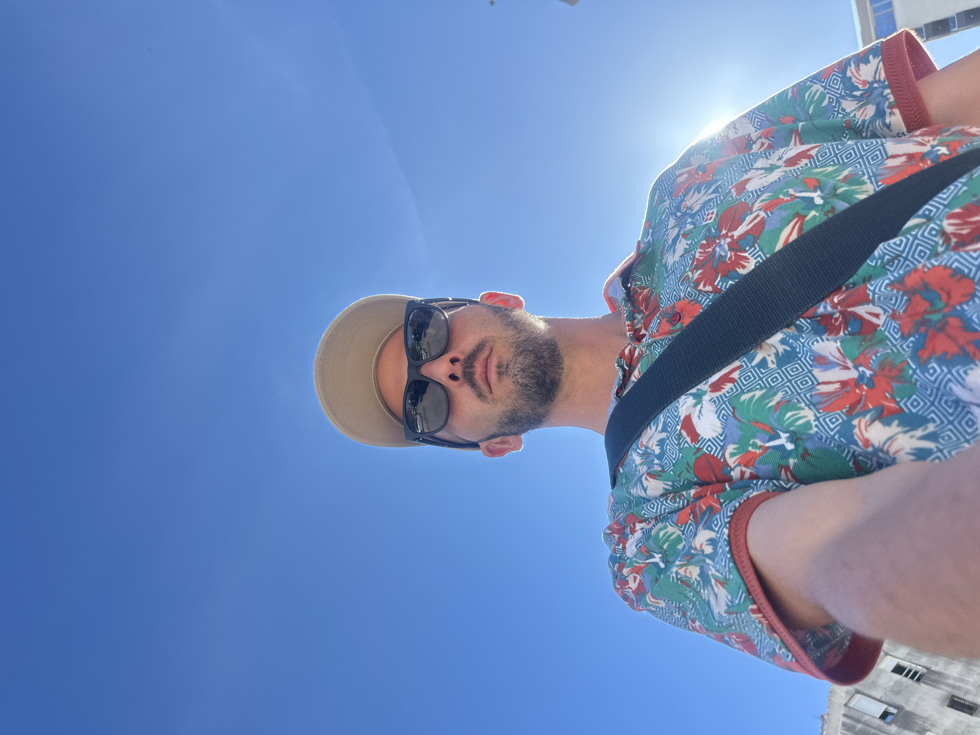 A low-angle selfie captures a man outdoors in Budva, Montenegro, under a clear blue sky. He directly faces the camera, wearing dark sunglasses, a beige baseball cap, and a vibrant short-sleeved collared shirt featuring a red, green, blue, and white tropical/geometric pattern. A black strap from a bag crosses his chest. Strong direct sunlight from the upper right creates a prominent lens flare and illuminates the right side of his face and body. To the left, a portion of a light-colored, multi-story building with several windows is visible. A faint outline of another structure is discernible on the far right edge of the frame.