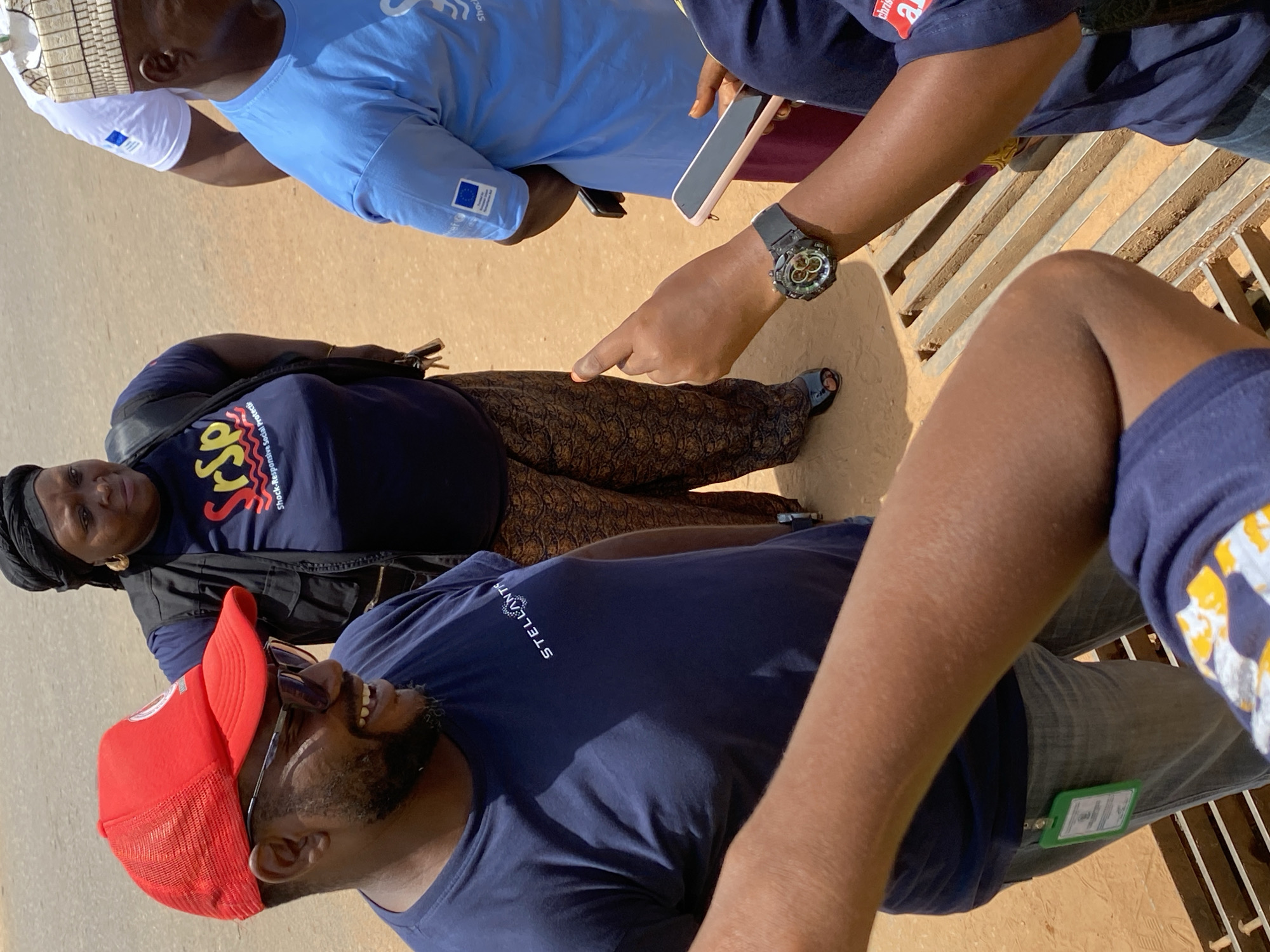 A group of four individuals are outdoors on a sunny day in what appears to be Sabon Tasha, Nigeria. The ground is light brown, possibly sandy or dusty. A Black woman is wearing a dark blue t-shirt with colorful writing and a black head covering, and a black vest. A Black man wearing a red cap and sunglasses is smiling. Another Black man is wearing a light blue t-shirt with a European Union logo. A fourth Black man wears a dark blue t-shirt and jeans. A black wristwatch is visible on one of the man's wrists. A hand is pointing toward a section of metal grating. A green identification badge is attached to the jeans. A mobile phone is also visible.