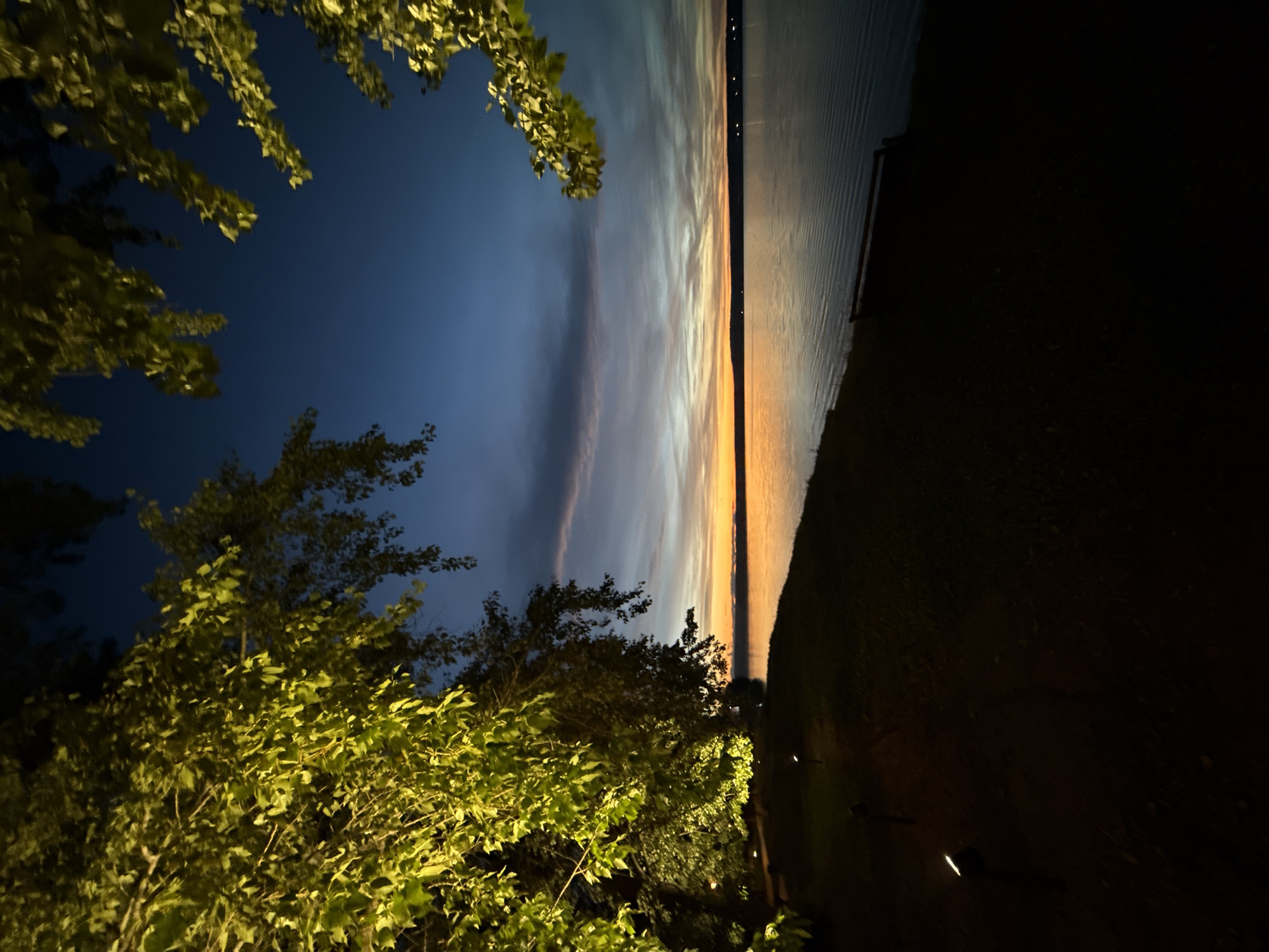 The image depicts a serene waterside scene at dusk, likely in Votkinsk, Russia. The sky transitions from a deep blue at the top to hues of orange and pink near the horizon, indicating the setting sun. A body of water, possibly a lake or river, fills the right side of the frame, reflecting the soft light of the sky. The shoreline in the foreground is dimly lit, with a path and small landscape lights visible. Lush green foliage, illuminated by artificial lights, drapes from the upper left and right corners of the image, framing the twilight sky and water. The overall atmosphere is tranquil and peaceful.