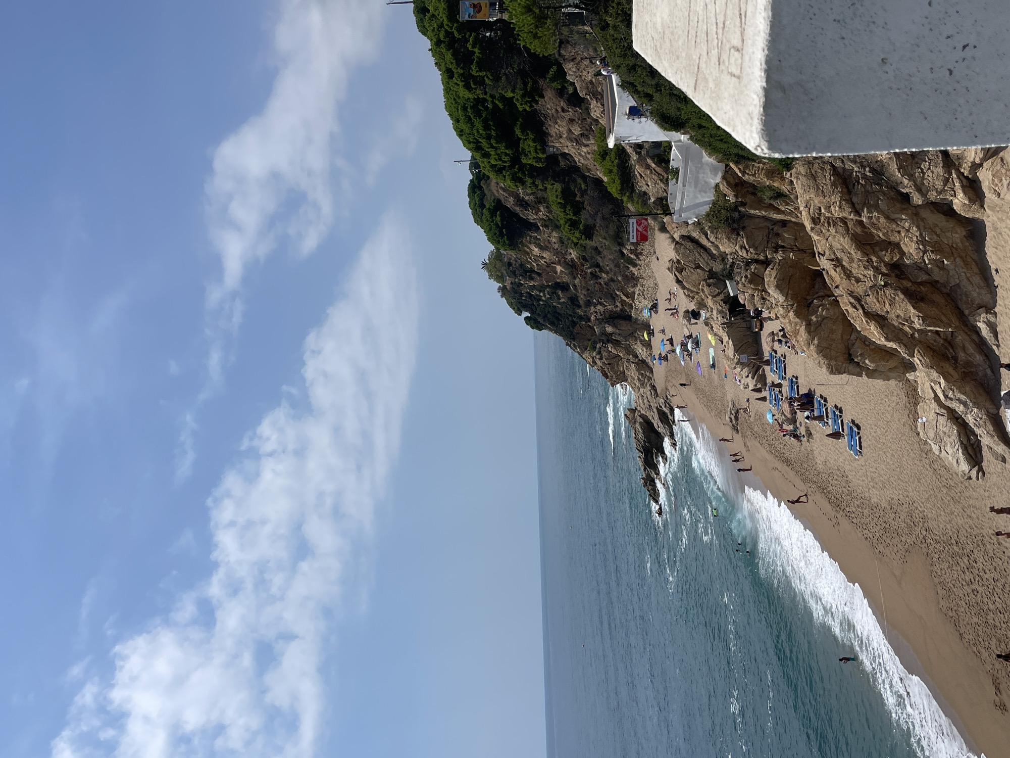 The image shows a scenic view of a crowded beach with people sunbathing and swimming in the ocean. The beach is situated at the base of a rocky, tree-covered cliffside. There are several beach chairs and umbrellas set up on the sand. A building with white walls and green foliage is visible on the cliffside. The ocean water is a deep blue, and the sky is clear with a few white clouds. The location is identified as a beach in Calella, Spain, likely with a view from a higher vantage point such as a cliffside restaurant or viewpoint.