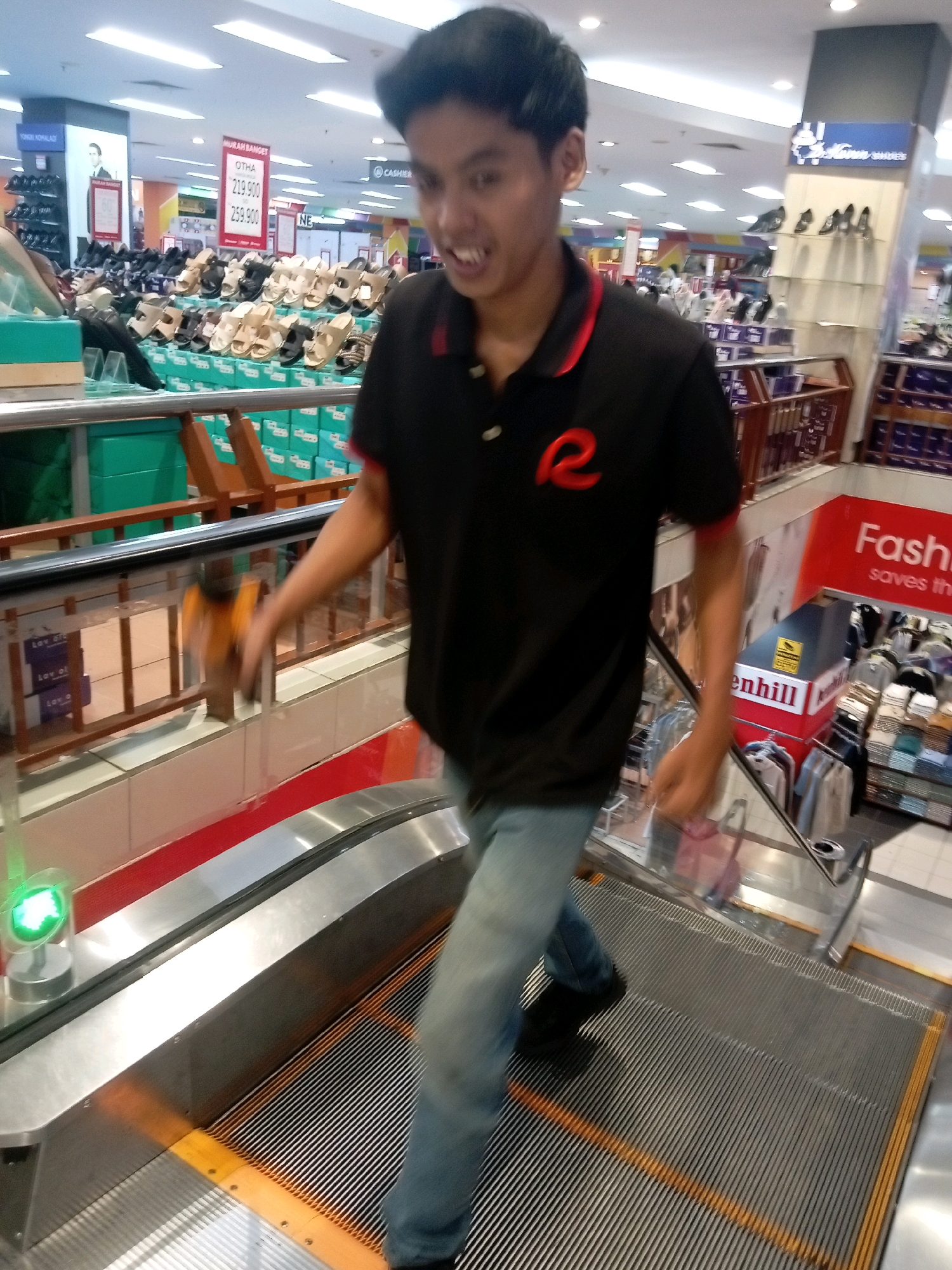 A young Indonesian male with dark hair and wearing a black polo shirt with a red logo is standing on an escalator in a shopping mall in Batam, Indonesia. The man is holding an orange object in his right hand. The escalator is moving upwards, and the man is looking towards the viewer. In the background, there are shelves stocked with shoes and other merchandise, as well as signs advertising sales. The lighting in the mall is bright, with fluorescent lights overhead. The scene appears to be captured during daytime.