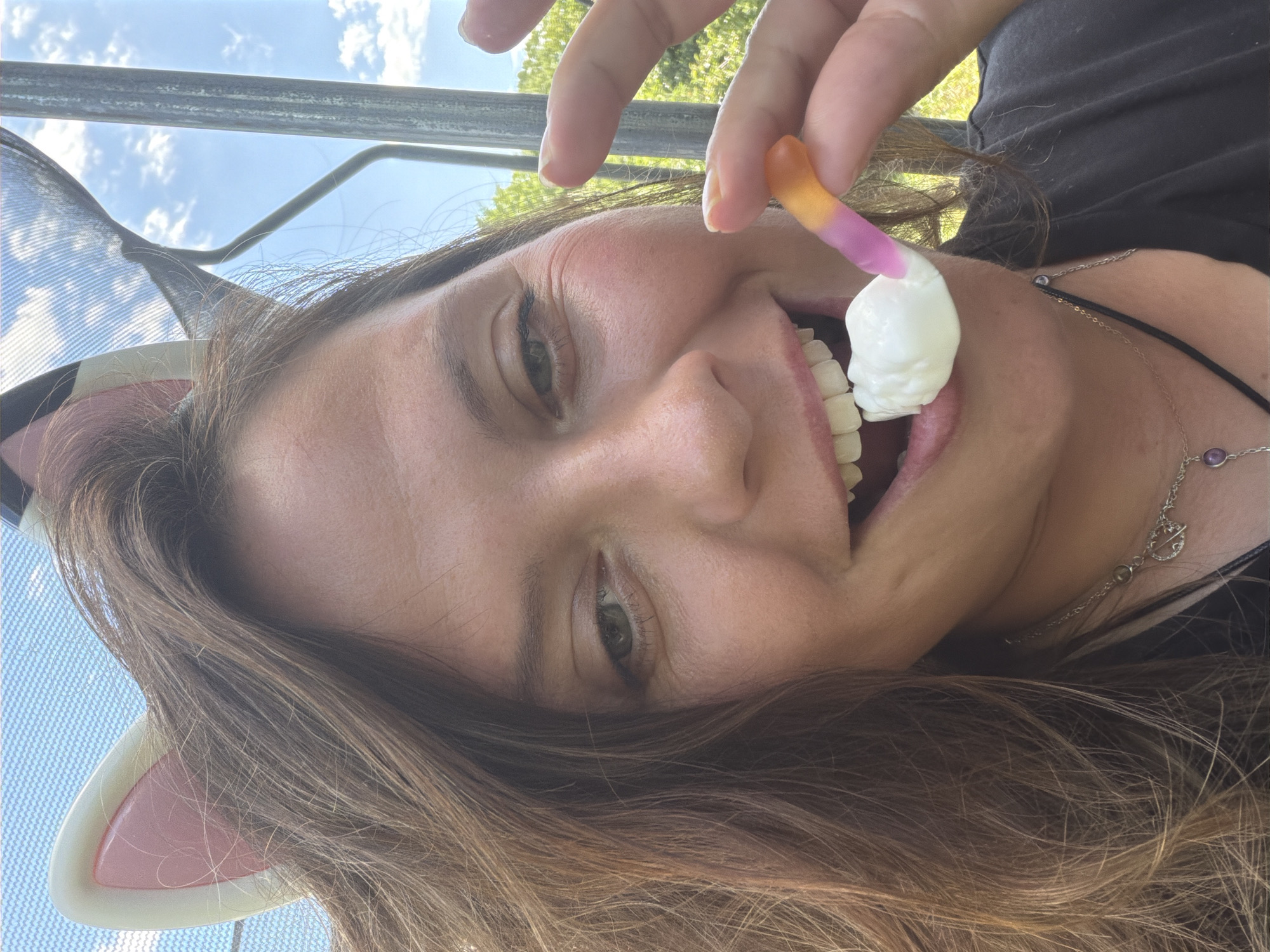 The media file shows a close-up, upside-down shot of a person's face as they are about to eat a gummy worm and a white, skull-shaped candy. The person has brown hair and is wearing cat-ear headwear. The background suggests an outdoor setting with a bright sky and some greenery, possibly in Kamianka-Buzka, Ukraine. The lighting indicates it is daytime. The person appears to be smiling and in a playful mood.