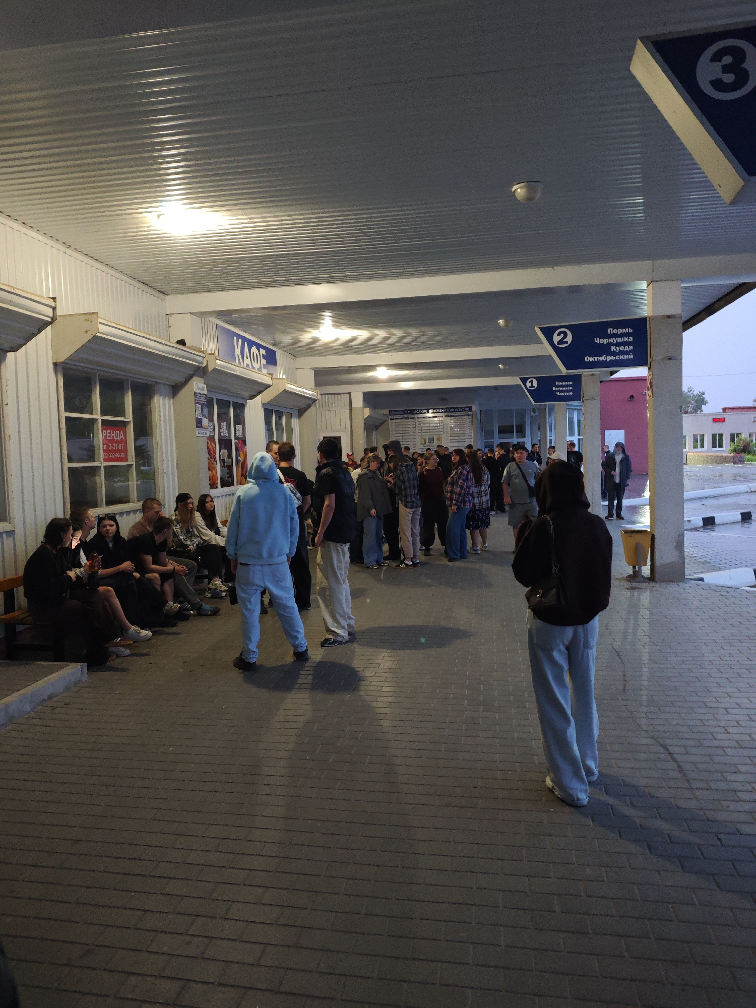 A group of people are standing and sitting in an outdoor covered area, likely a bus station or waiting area. The architecture suggests a functional, perhaps slightly rundown, public space with a corrugated metal roof and a paved walkway. Several signs are visible, including one in Russian that reads "КАФЕ" (Cafe) and others indicating bus routes with destination names such as "Пермь," "Чернушка," "Куеда," "Октябрьский," "Ижевск," "Воткинск," and "Частые." The lighting is artificial, casting shadows on the ground, indicating it's either early morning or evening. Some individuals are talking amongst themselves, while others appear to be waiting or observing. The overall impression is of a temporary pause in a journey or a gathering point before departure. The presence of Russian language signage indicates the location is in Russia. The specific destination names on the signs (e.g., Perm, Chaikovsky) strongly suggest this is the bus station in the city of Chaikovsky, Russia, as these are common routes originating or passing through the region.