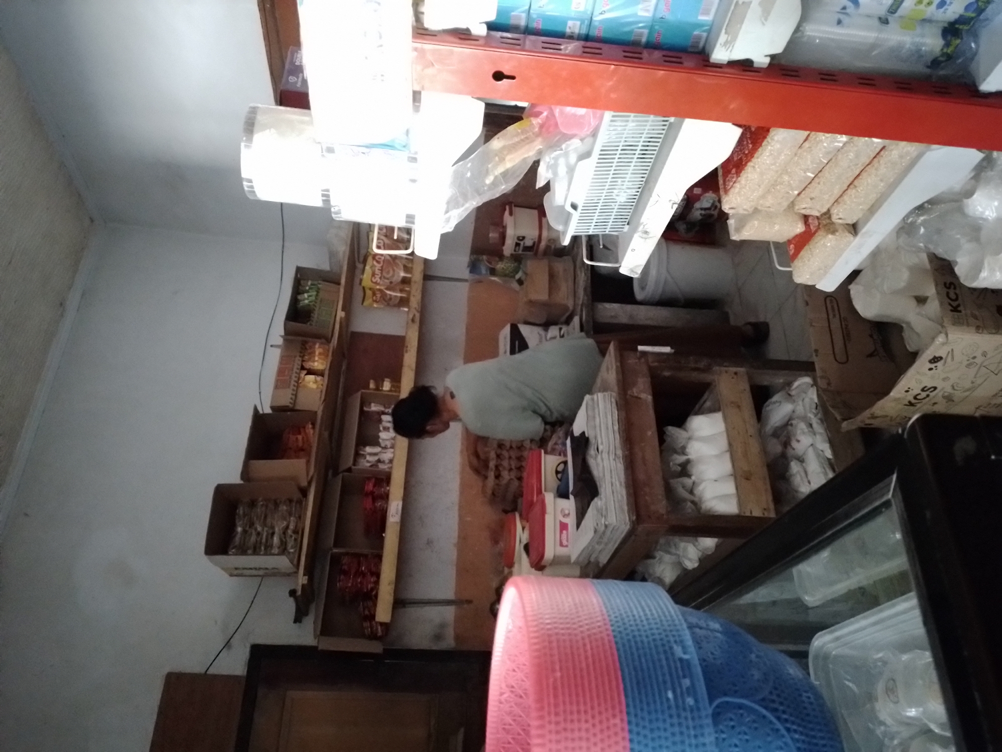 The image shows the interior of a small shop, likely a grocery or convenience store, in Ngancar, Indonesia. In the center of the frame, a woman wearing a light green t-shirt is standing and facing away from the camera, sorting through a stack of egg cartons on a wooden table. To the left of the woman, there is a stack of blue and pink plastic baskets. Shelves above the woman are stocked with various products in boxes and bags, including what appear to be packaged food items and cleaning supplies. A refrigeration unit with glass doors is partially visible in the lower left corner. To the right, a red shelving unit displays packaged goods and containers. The environment appears somewhat cluttered, suggesting a busy retail space.
