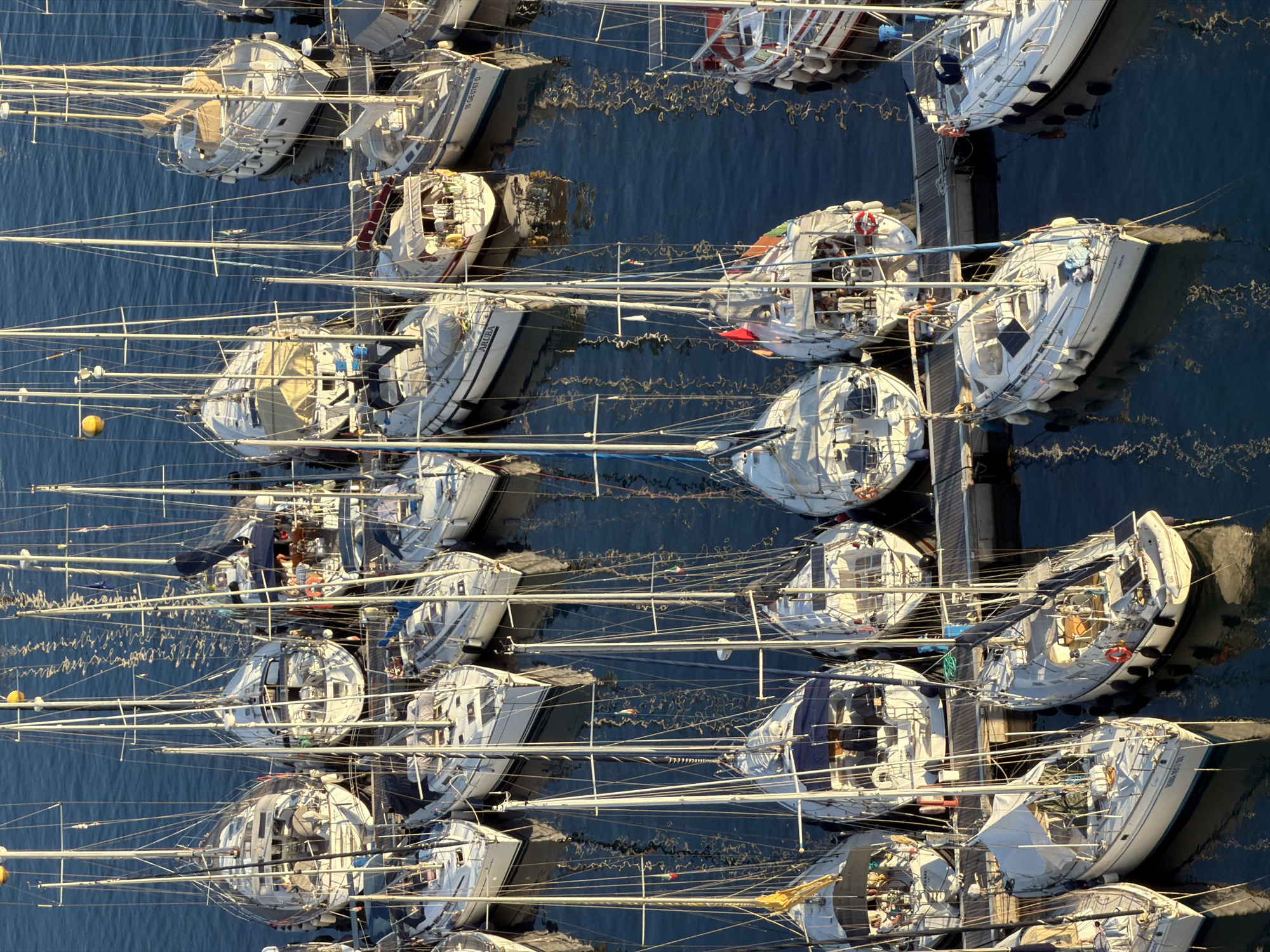This is an aerial, high-angle, full shot of a marina in Santa Maria Navarrese, Italy, filled with numerous sailboats docked in calm, blue water. The sun is casting long shadows, indicating it is either early morning or late afternoon. The boats are white with various colored accents and sails, some furled and some loosely draped. Several boats have protective covers over their booms and cockpits. Some boats are docked side-by-side, while others are moored individually. A wooden dock runs along the bottom of the frame, with a few people visible on the boats, appearing relaxed and engaged in conversation or tending to the boats. One boat in the foreground has a license plate that reads "15 GE 0167 D". The water reflects the sky and the boats, creating a shimmering effect. The overall scene conveys a sense of tranquility and leisure associated with sailing.