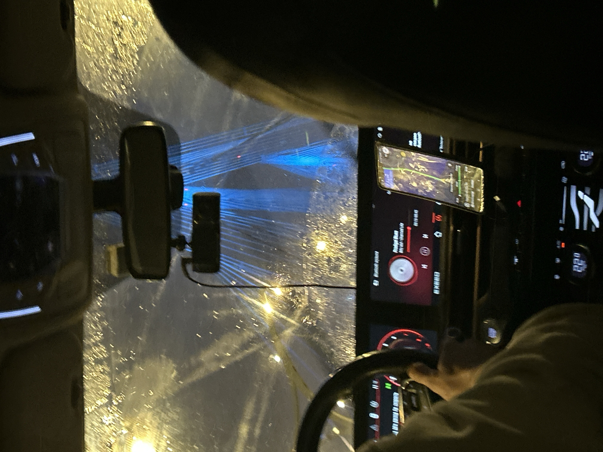 Here's a description of the image:
The image depicts the interior of a car at night, likely during rainfall. The windshield is covered with water droplets and reflects the city lights of Saint Petersburg, Russia. Visible are distinctive blue light beams, a feature of the city's bridges. The rearview mirror is centrally located, with a small electronic device attached. The dashboard features a large touchscreen display showing navigation and music controls. Another screen to the right shows vehicle data. A hand is visible on the steering wheel, indicating that someone is driving. The person's arm is clad in a light-colored garment.