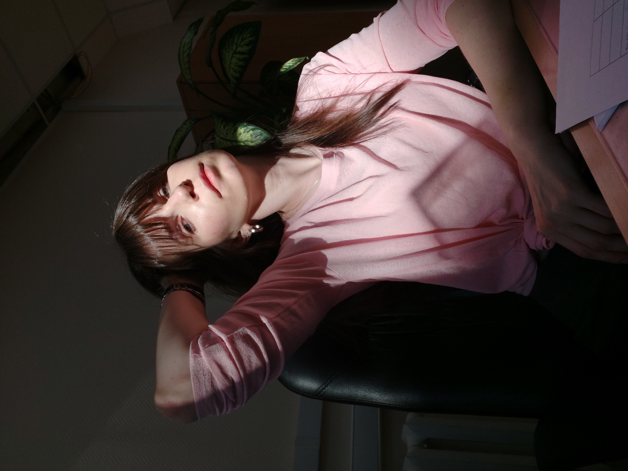 The image shows a woman with brown hair and fair skin. She is wearing a pink long-sleeved shirt and is sitting in a black chair. Her left arm rests on the chair while her right arm is bent. There is a potted plant with green leaves behind her head. The background is dark with a beam of light shining on her face and shirt. There is a piece of paper visible on the right side of the image. The location is possibly Chaikovsky, Russia.