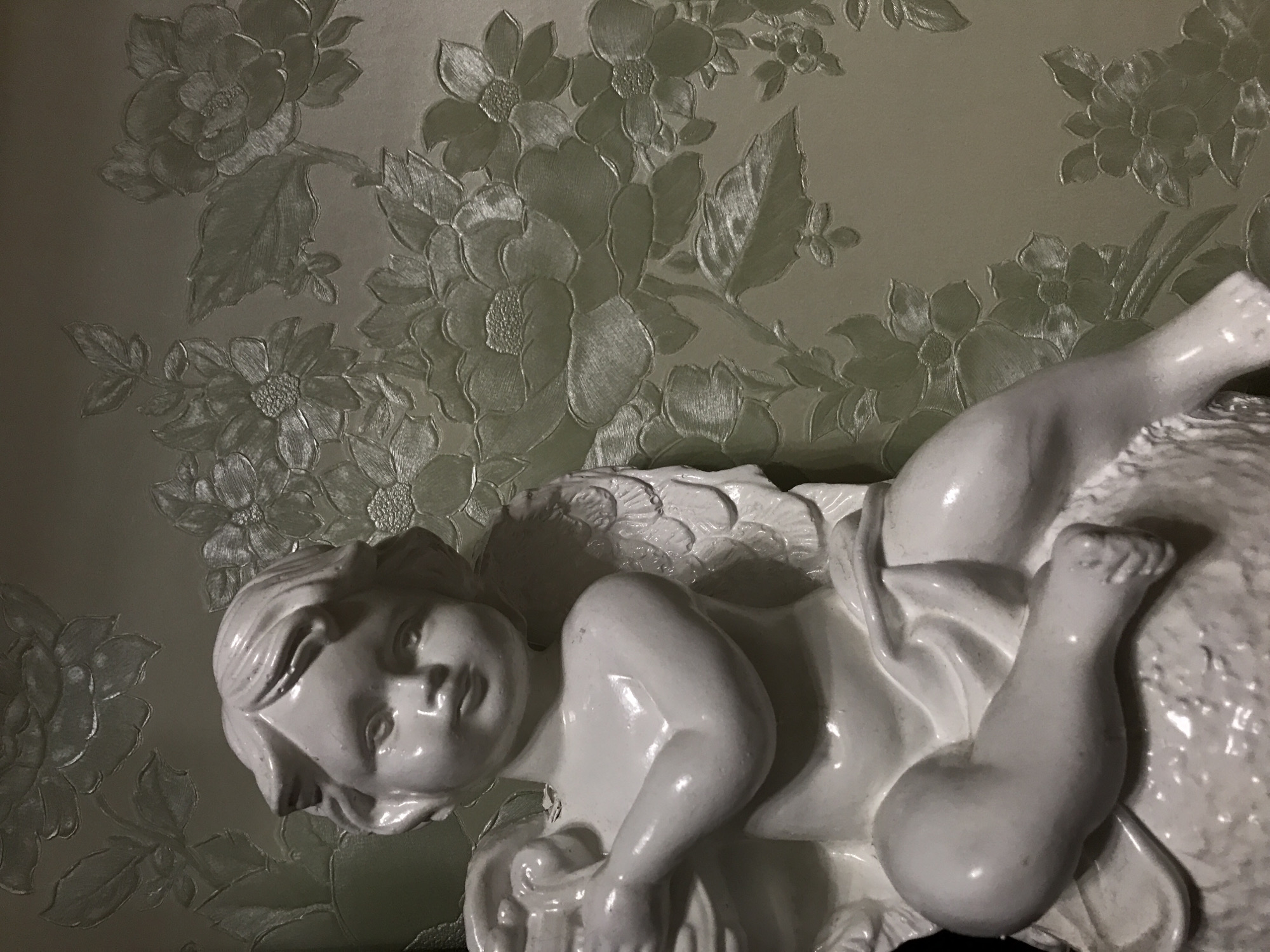 A close-up, slightly angled shot depicts a white ceramic angel figurine against a floral patterned wall. The angel lies prone, with its head to the left and legs extended to the upper right. The figurine is detailed, showing features of the face, wings, and drapery. The wall behind the figurine features a raised floral pattern in a pale color.
There is no verifiable evidence to suggest this image was taken in Perm, Russia.