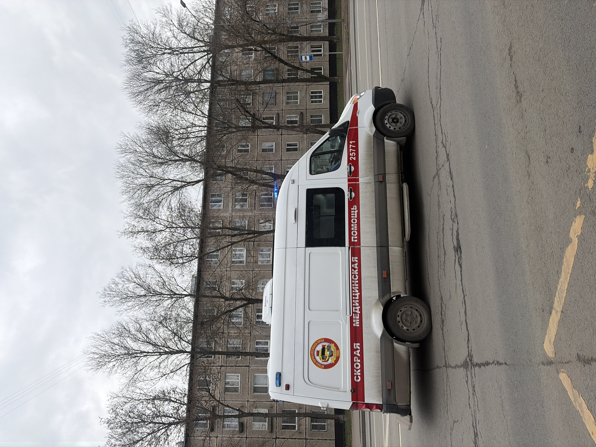 The image depicts a white and red ambulance parked on a city street. The ambulance has the words "СКОРАЯ МЕДИЦИНСКАЯ ПОМОЩЬ" written on its side, which translates to "Emergency Medical Assistance." There is also a circular emblem with a yellow ambulance inside. The number 25771 is visible on the side.
The ambulance is parked next to a gray apartment building. Bare trees are visible in front of the building. The street is cracked and marked with yellow paint.
The scene appears to be in a residential area in the city of Saint Petersburg, Russia.
