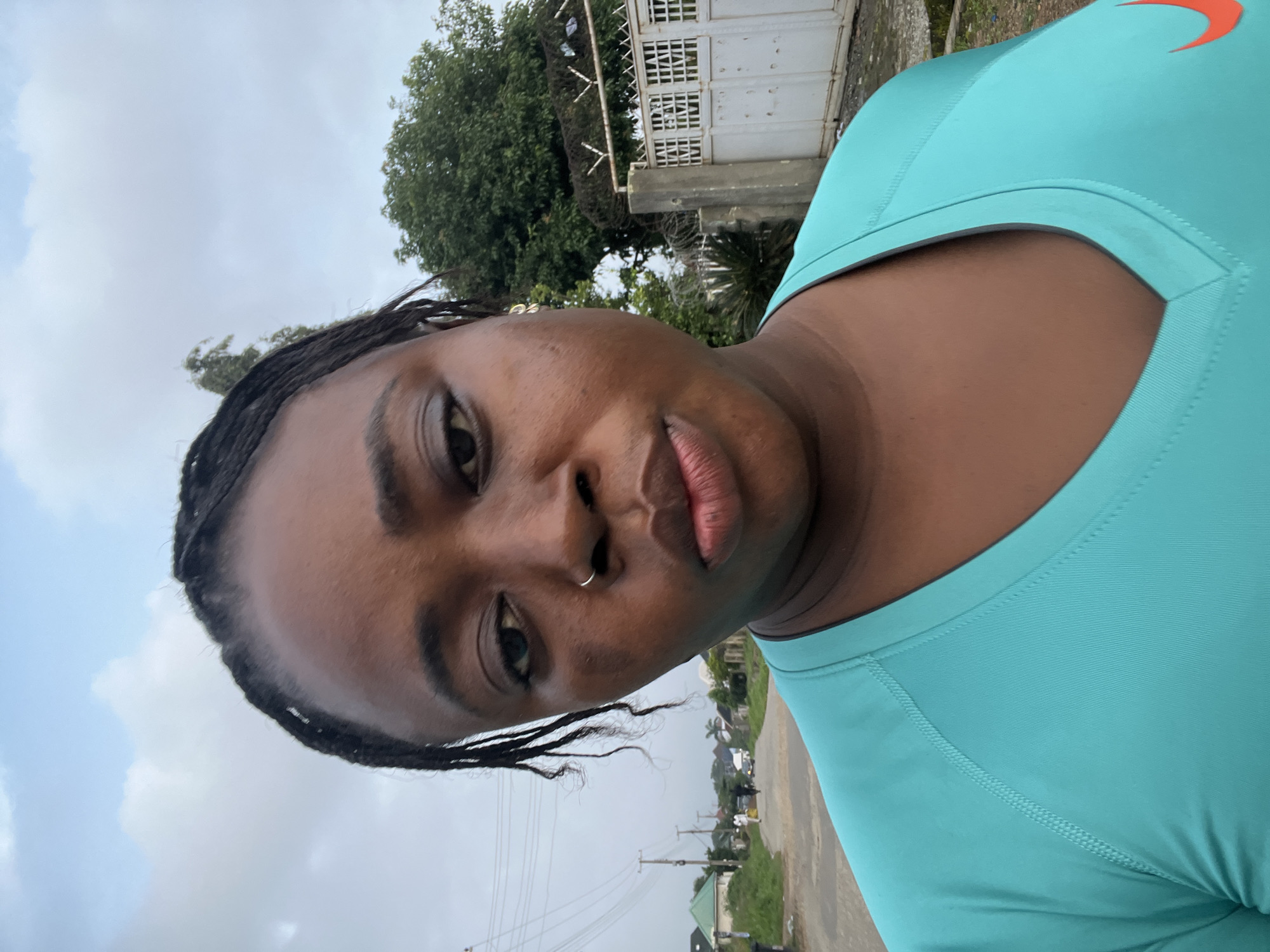 A medium shot captures a Black woman posing for a selfie outdoors in Abuja, Nigeria. The woman is positioned centrally in the frame, facing the camera with a slightly pensive expression. She has dark, braided hair and wears a turquoise V-neck t-shirt with a subtle orange Nike logo visible on the right side of her chest. A silver nose ring adorns her right nostril, and a small gold earring is visible on her right earlobe.
The background suggests a residential street. To the left, a paved road stretches into the distance, lined with houses and utility poles with visible power lines. Several people and a vehicle are faintly discernible further down the road. To the right, a white metal gate with intricate latticework is visible, behind which are trees and a wall topped with barbed wire. The sky above is overcast with clouds. The overall lighting suggests it is either early morning or late afternoon.